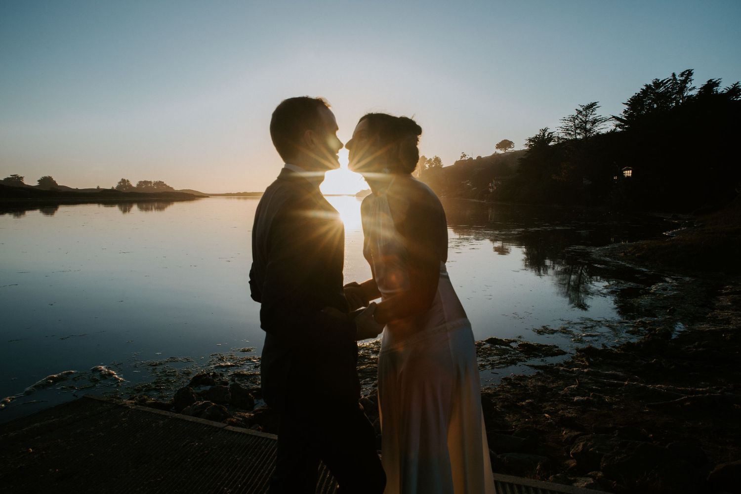 Silhouetted figures stand together against a glowing sunset sky reflected in calm waters at the shoreline.