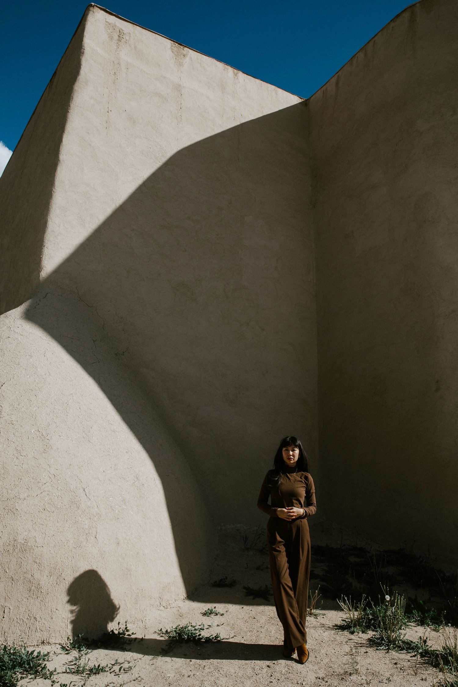 Woman in brown outfit standing against curved white stucco walls with dramatic shadows and blue sky.