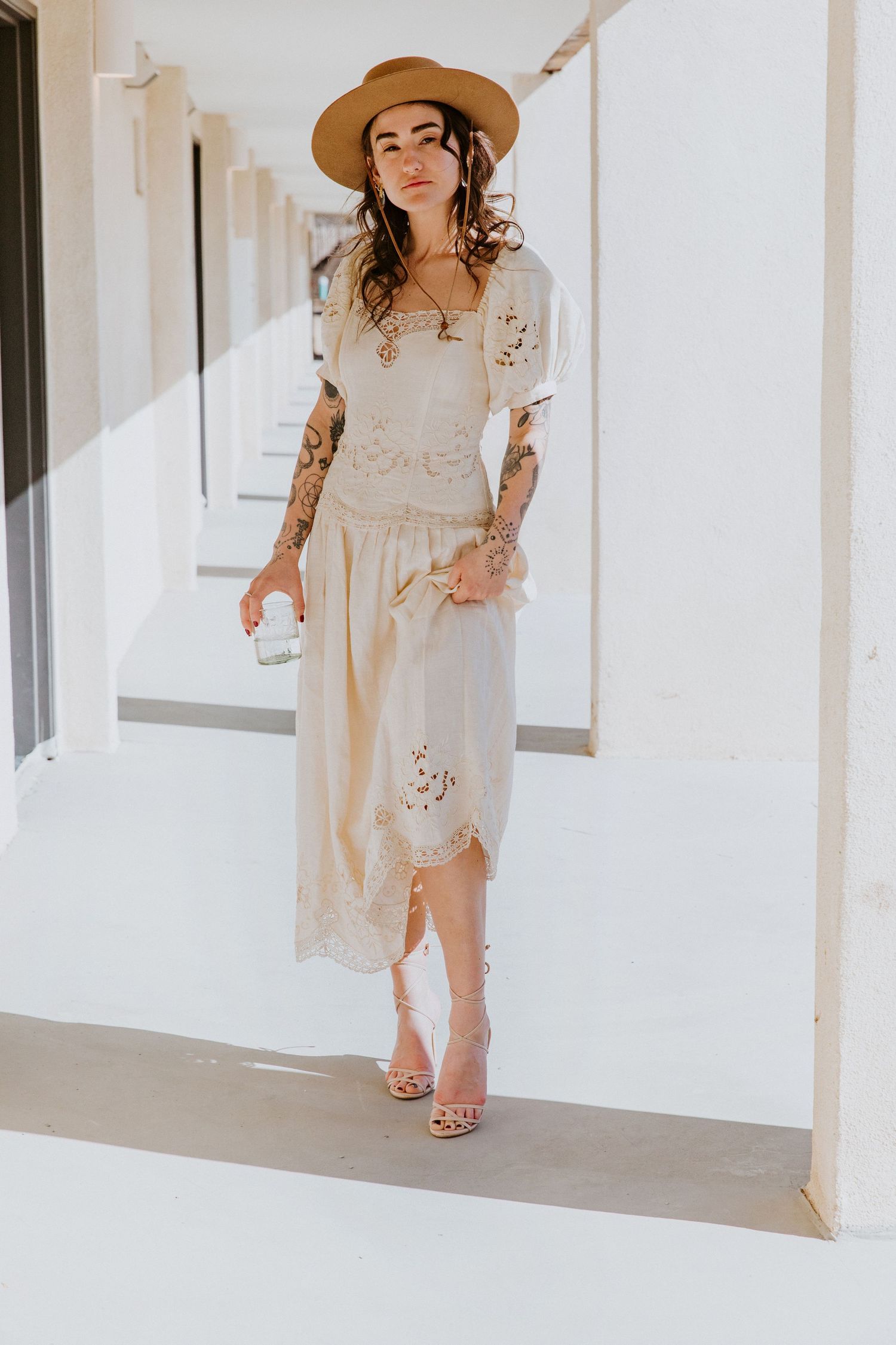 A figure in a flowing cream dress and sun hat walks through a bright white corridor.
