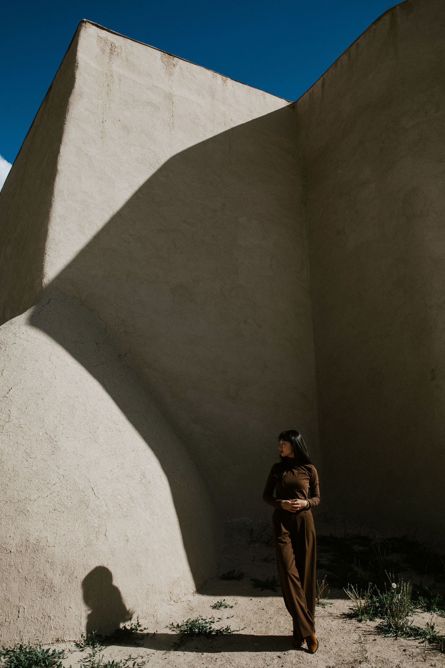 woman in brown jumpsuit standing against curved adobe wall with dramatic shadows and blue sky.