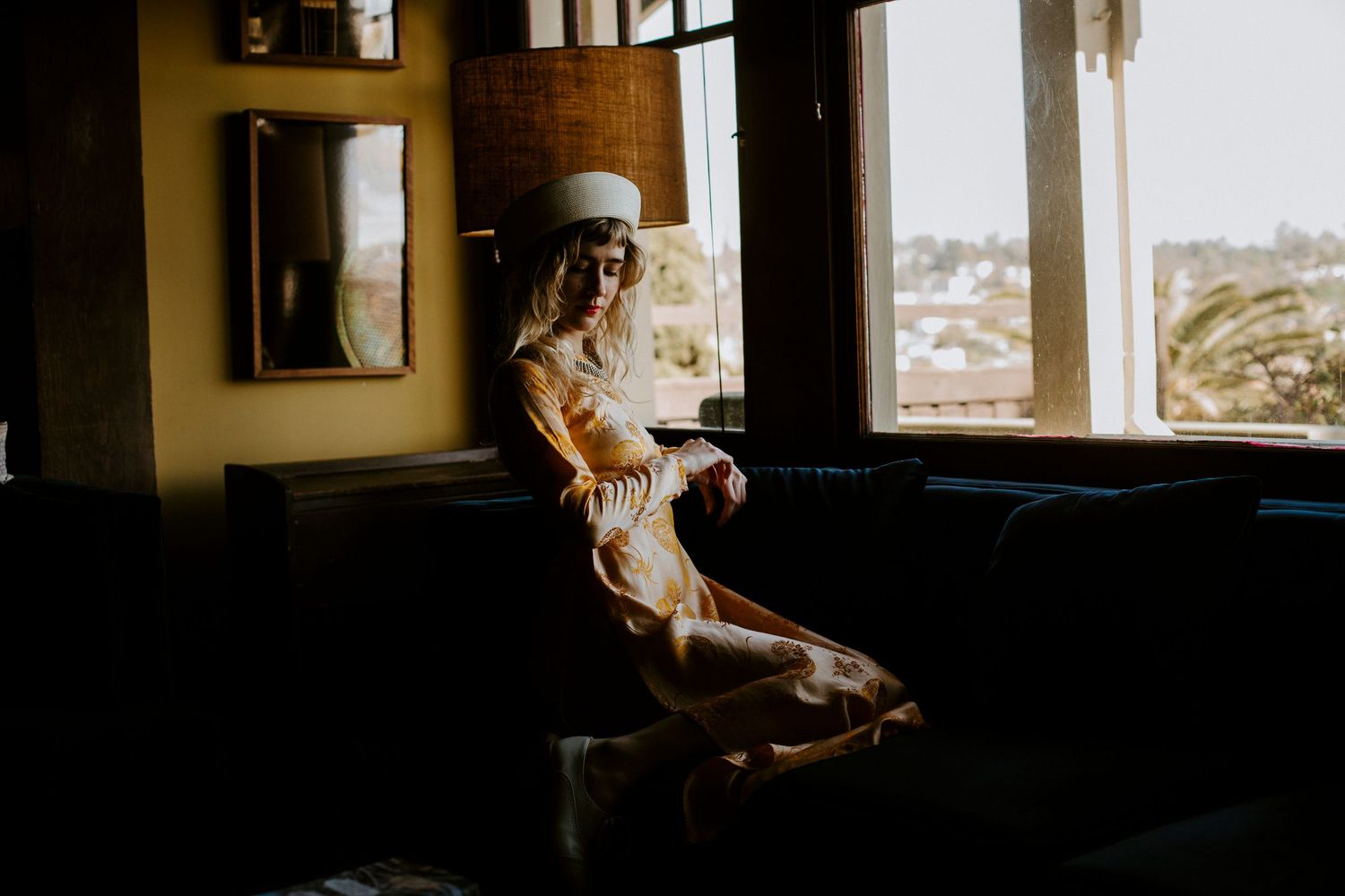 A figure sits by a large window in a dimly lit room, with warm sunlight casting atmospheric shadows across the interior.