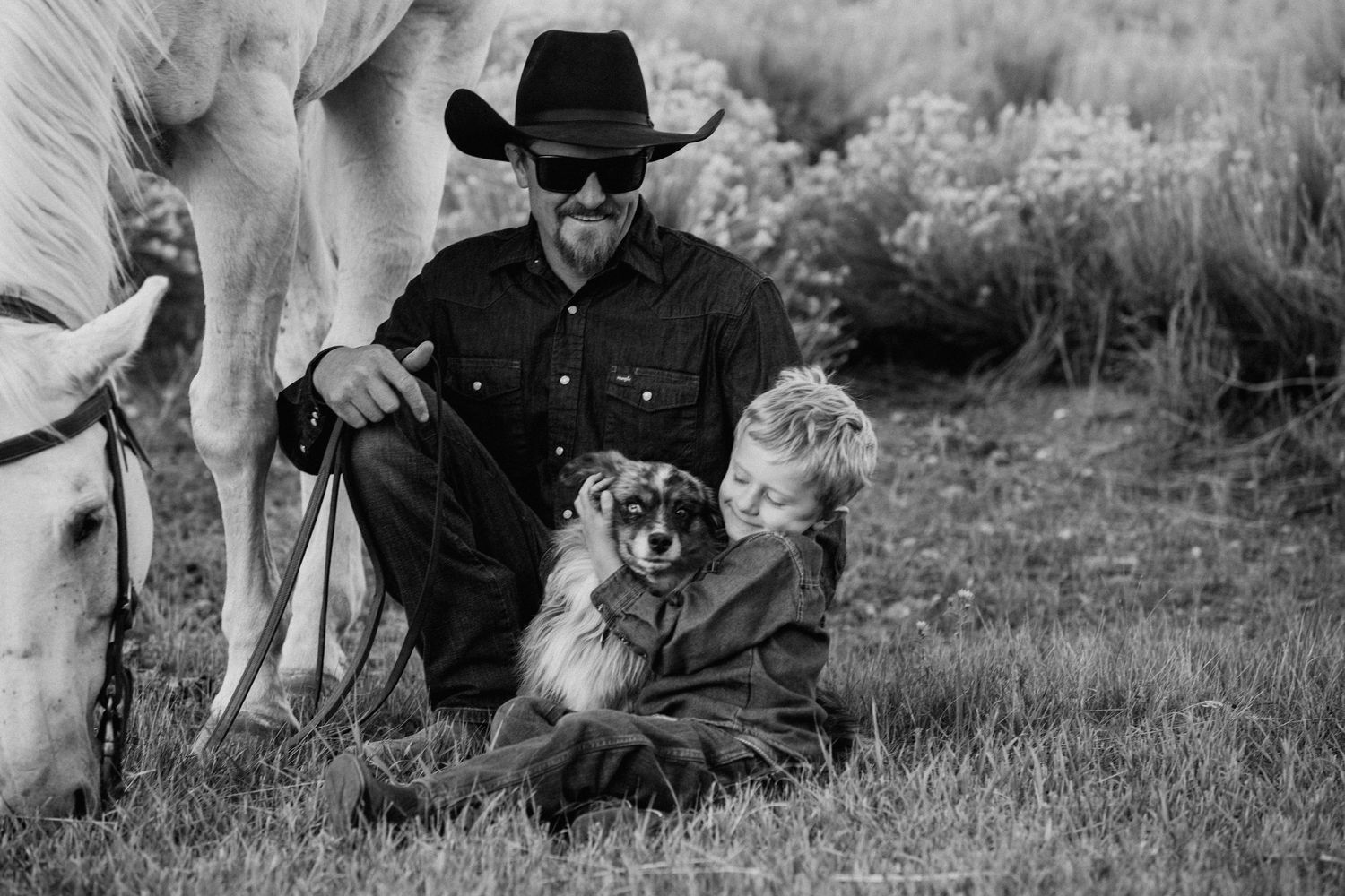 A black and white photo of a dog with two people sitting by a horse in a rural setting.