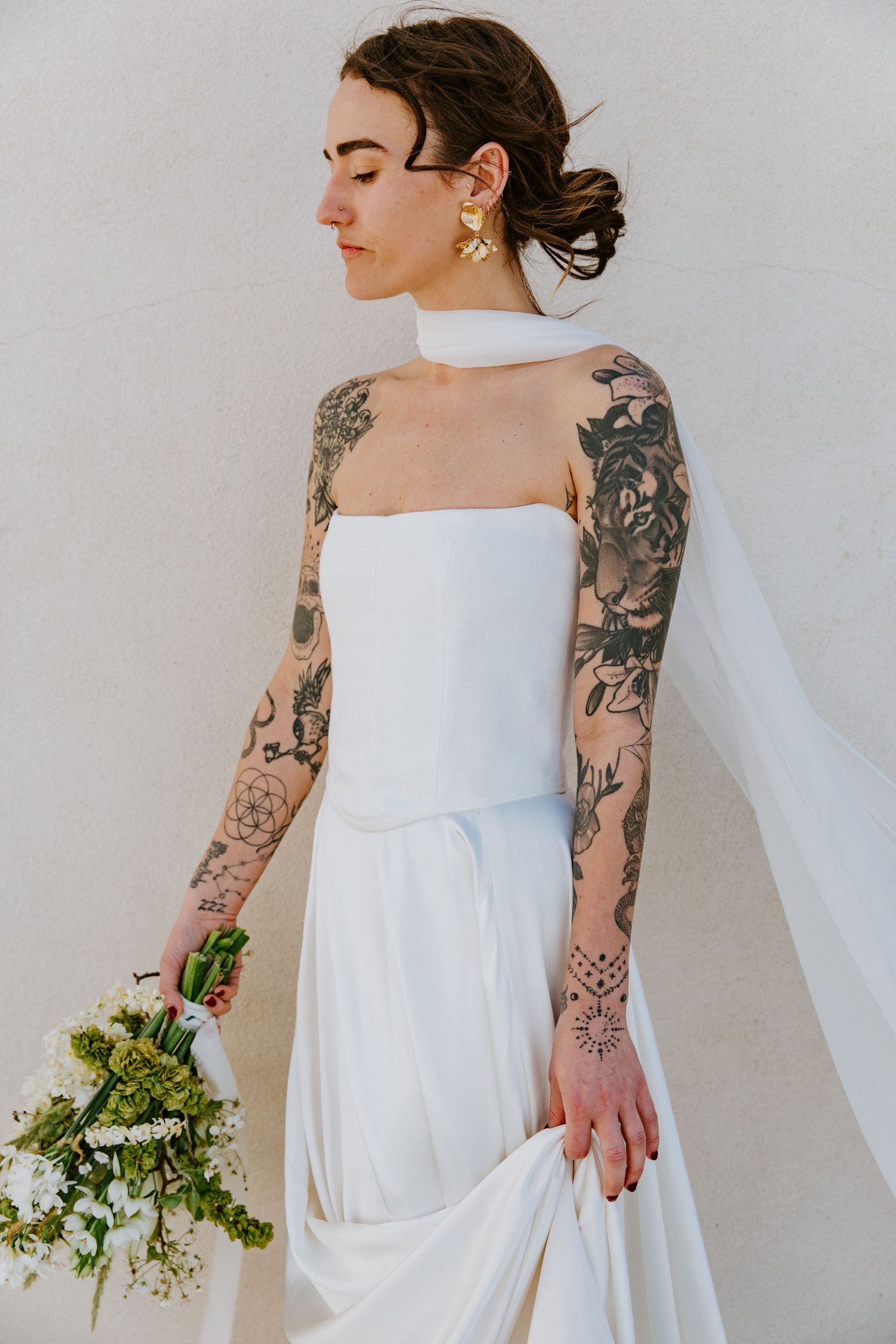 A bride in a modern white wedding dress shows off intricate sleeve tattoos while holding greenery against a light background.