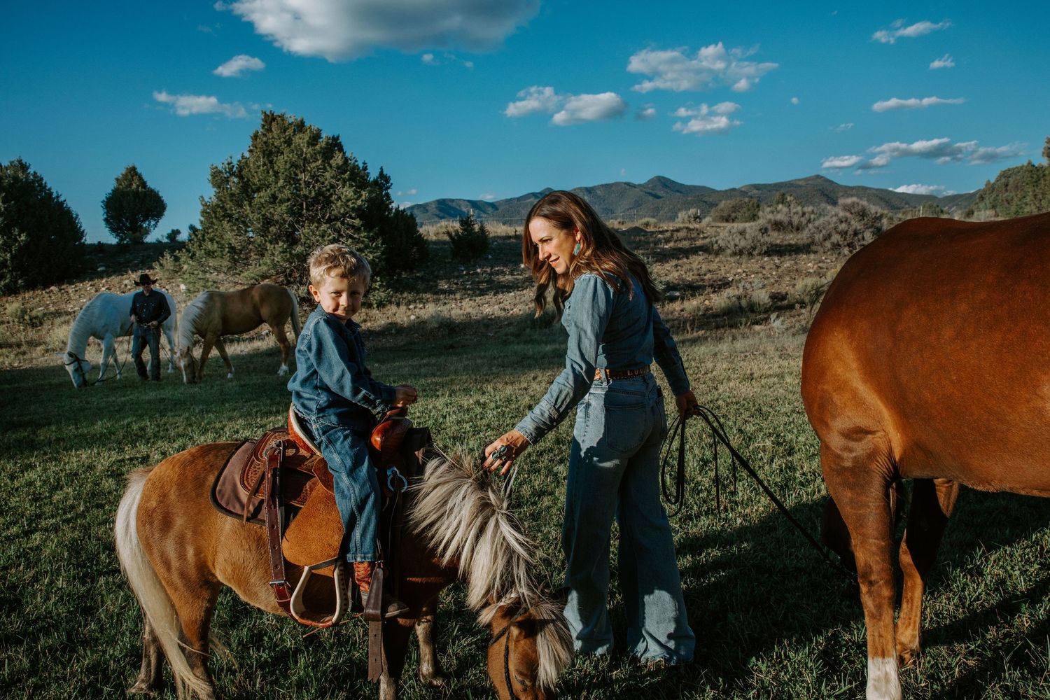 A person riding a pony in a scenic mountain landscape while another person stands nearby with horses grazing in the background.