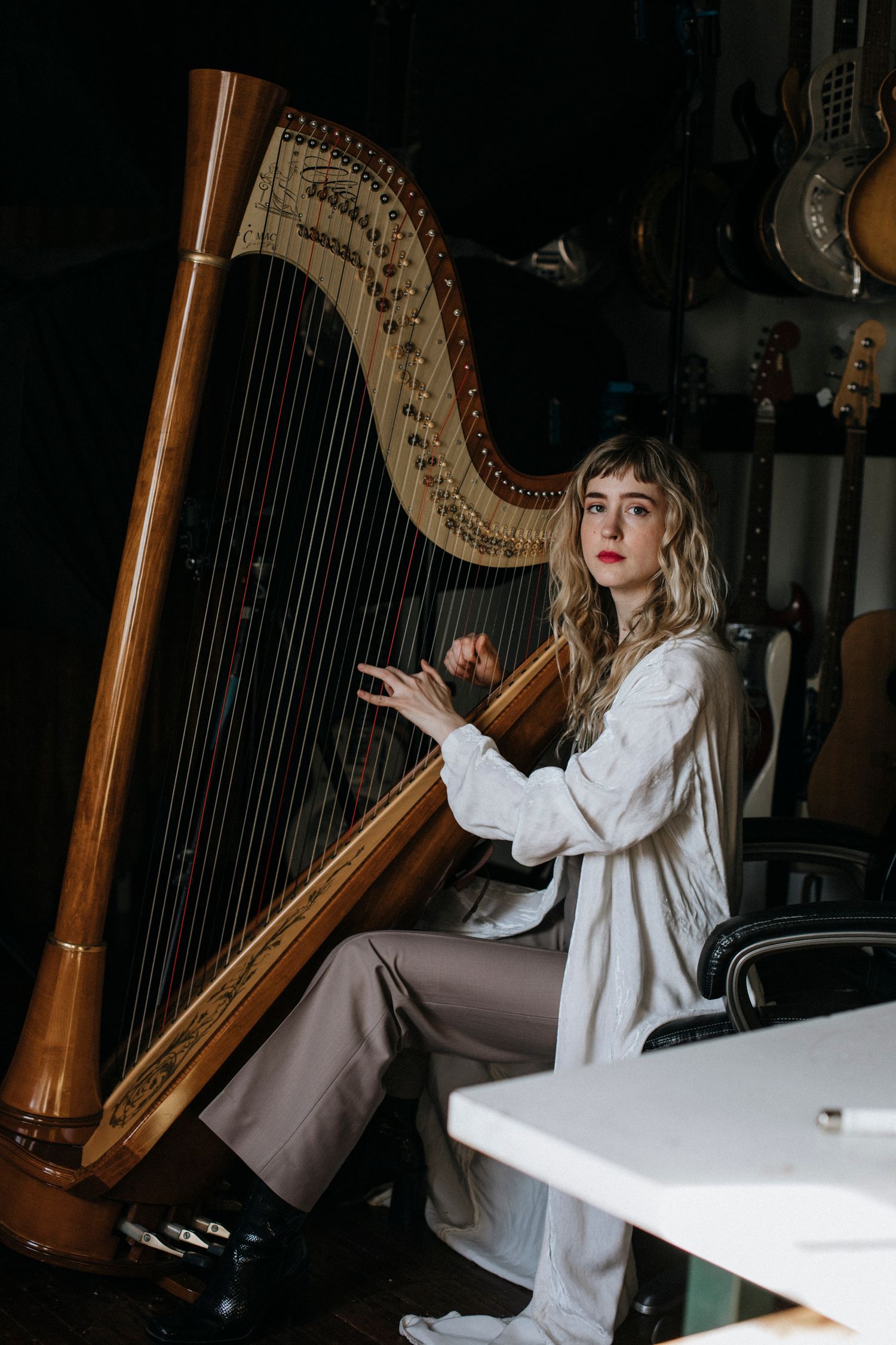 A musician sits at a grand pedal harp in a dimly lit room wearing a white flowing outfit.