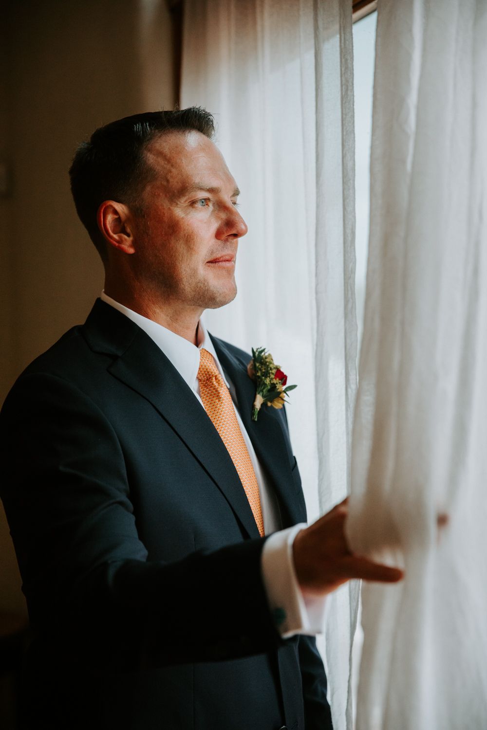 A groom in a dark suit with orange tie adjusts his boutonniere while standing by a window in natural light.