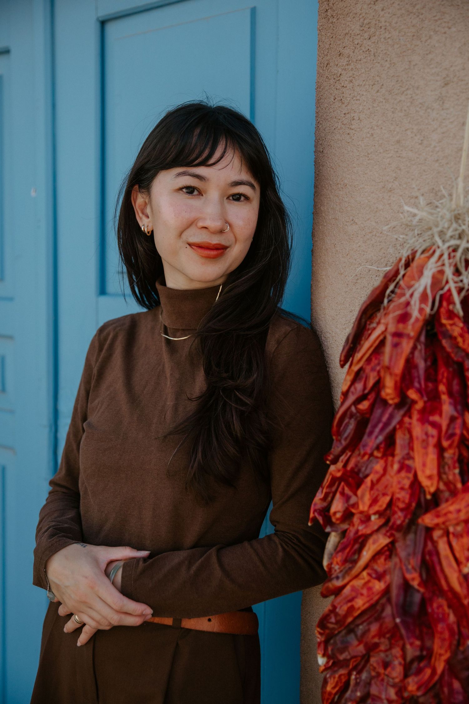 Woman in brown turtleneck poses beside hanging red chili peppers near a blue door and adobe wall.
