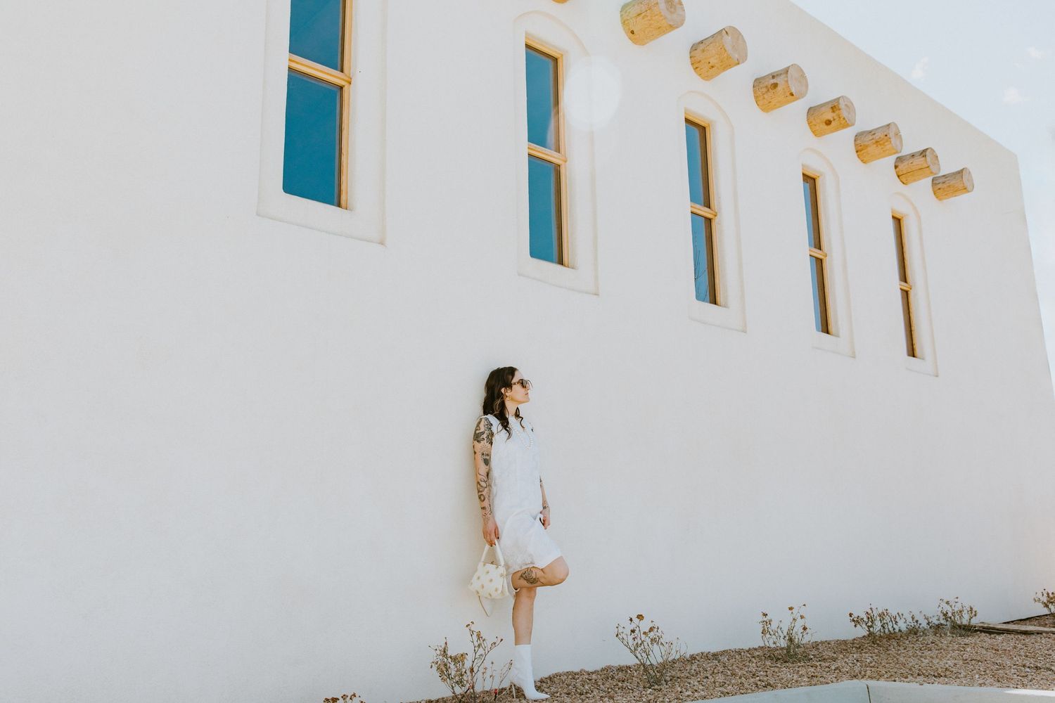 A person in a white dress stands against a stark white building with turquoise window frames in a minimalist setting.