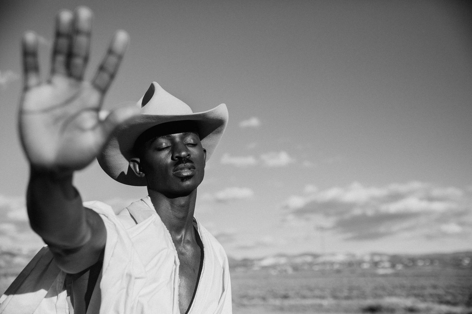 A person in a cowboy hat raises their hand against a dramatic black and white desert landscape.