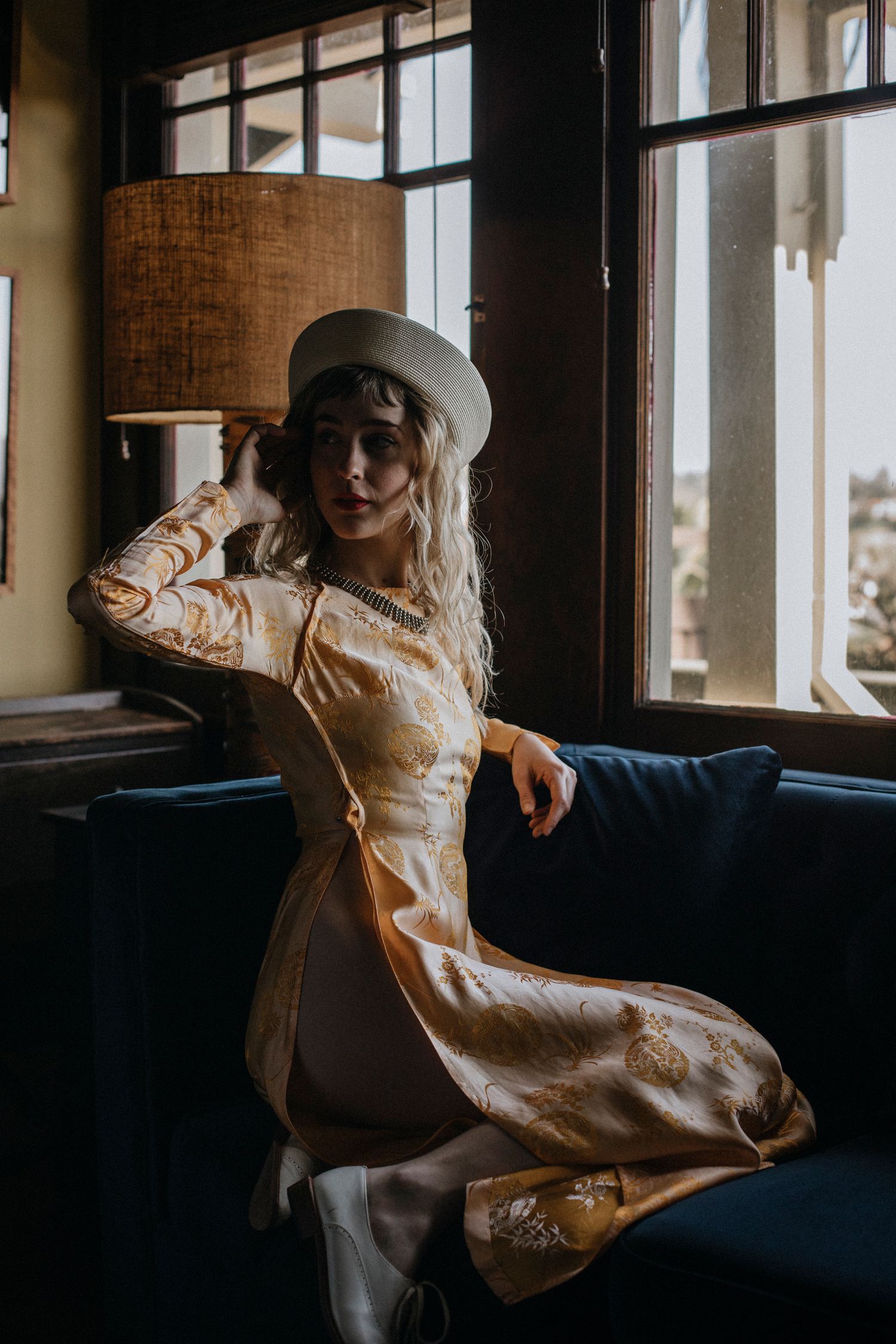A woman in a cream colored coat lounges on a blue couch near large windows in moody lighting.