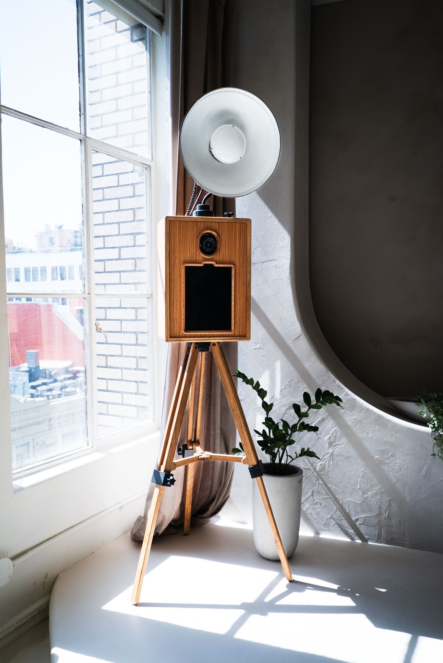 A vintage-style wooden speaker with tripod stand and white horn sits by a bright window in a minimalist room setting.