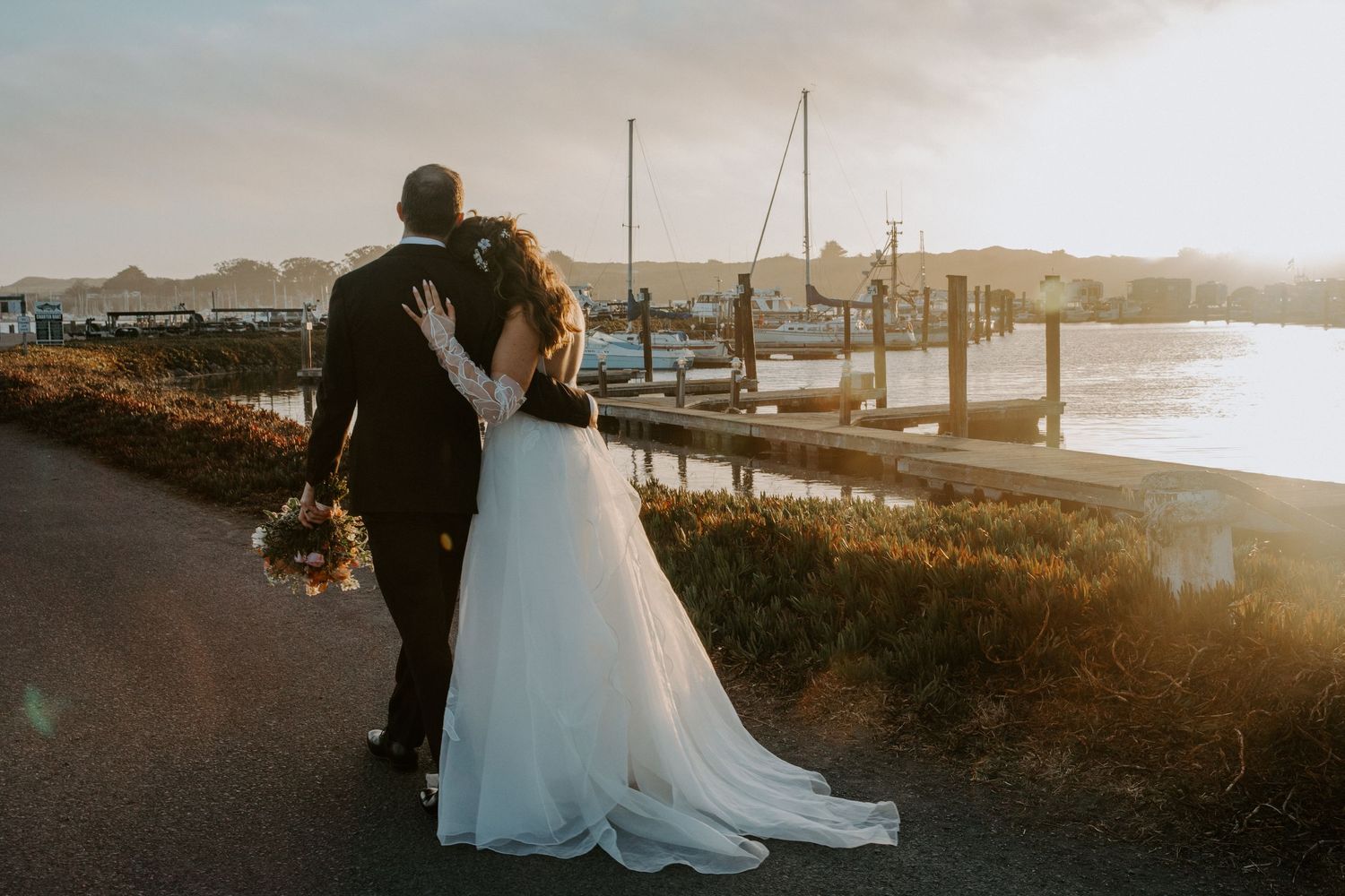 A couple shares an intimate moment at sunset by a marina with boats in the background.