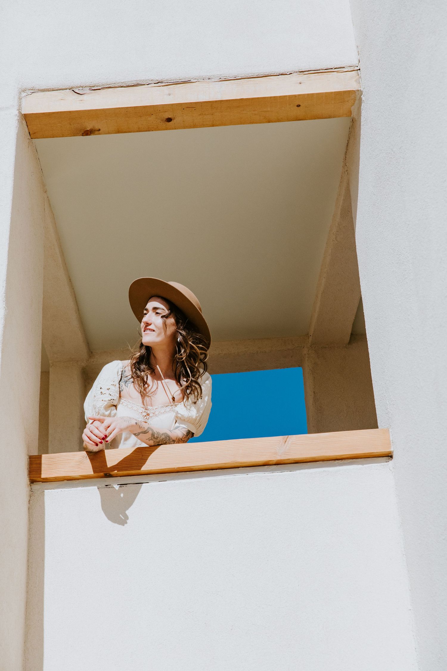 Person in a white dress and sun hat poses in an architectural space with white walls and rectangular openings.