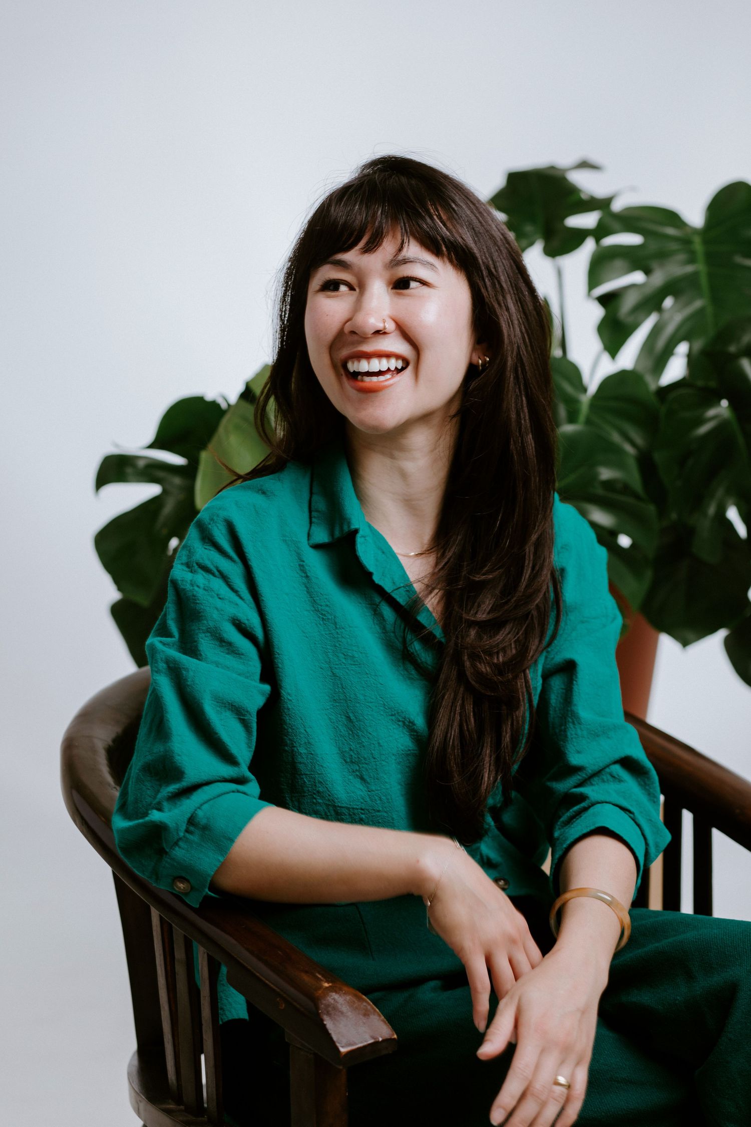 Smiling young woman in green linen shirt seated in wooden chair with monstera plant background.