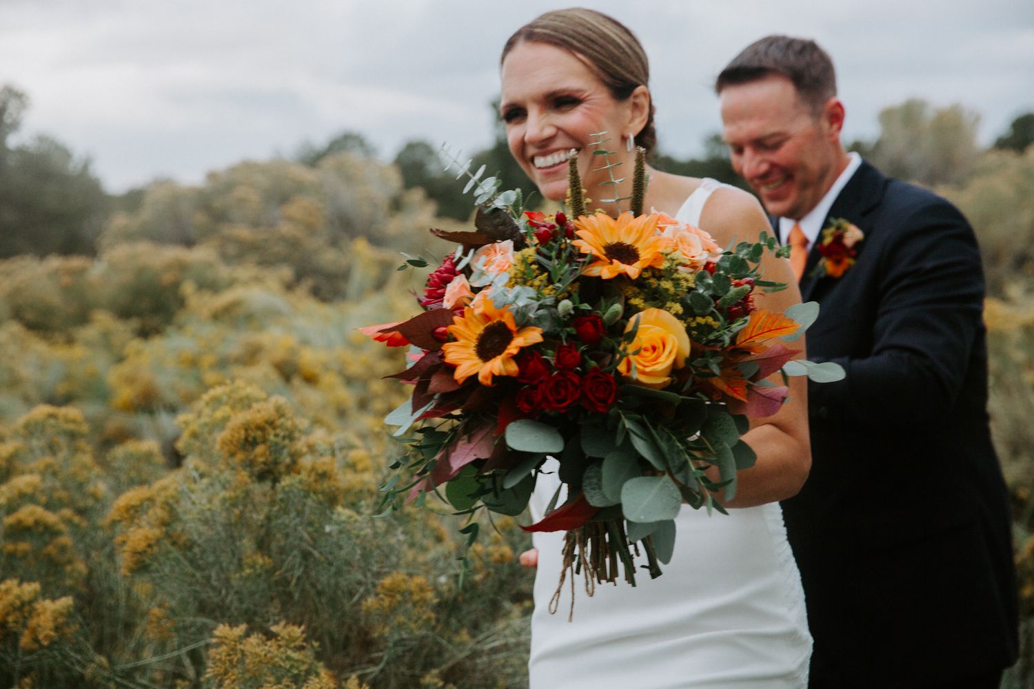 A couple shares a joyful moment in a wildflower field while holding a vibrant autumn bouquet with sunflowers and red blooms.