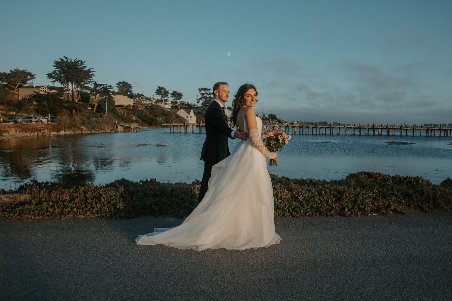 A beachside wedding couple poses at sunset with the ocean pier and rocky coastline in the background.