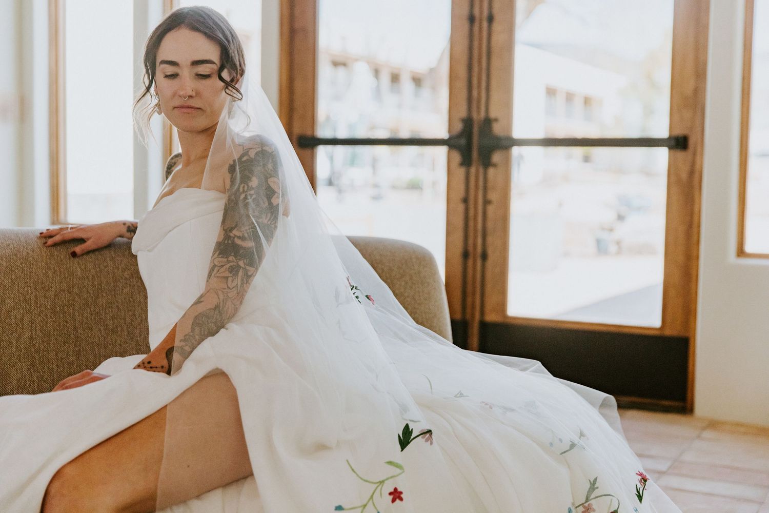A wedding dress hangs elegantly in a sunlit room with soft, romantic lighting and window reflections.