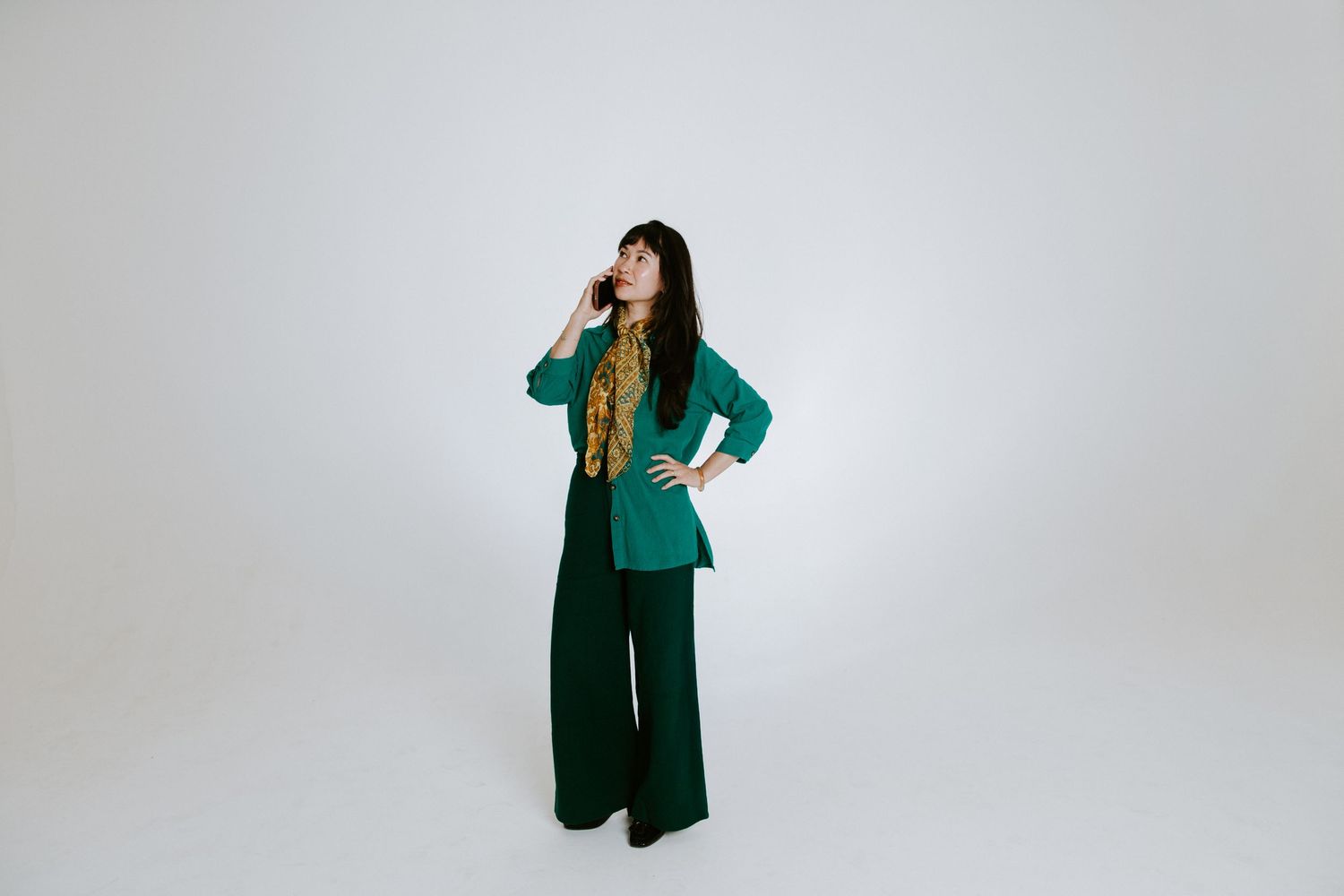 Woman in green wide-leg pantsuit with yellow scarf, posing thoughtfully against white background.