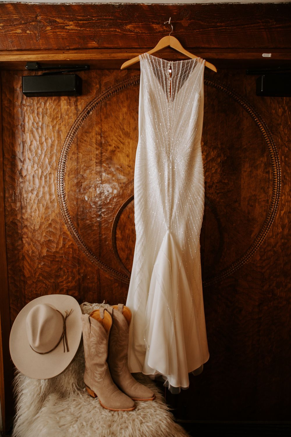 White sleeveless wedding dress hanging beside cowboy boots and hat against rustic wooden wall with carved details.