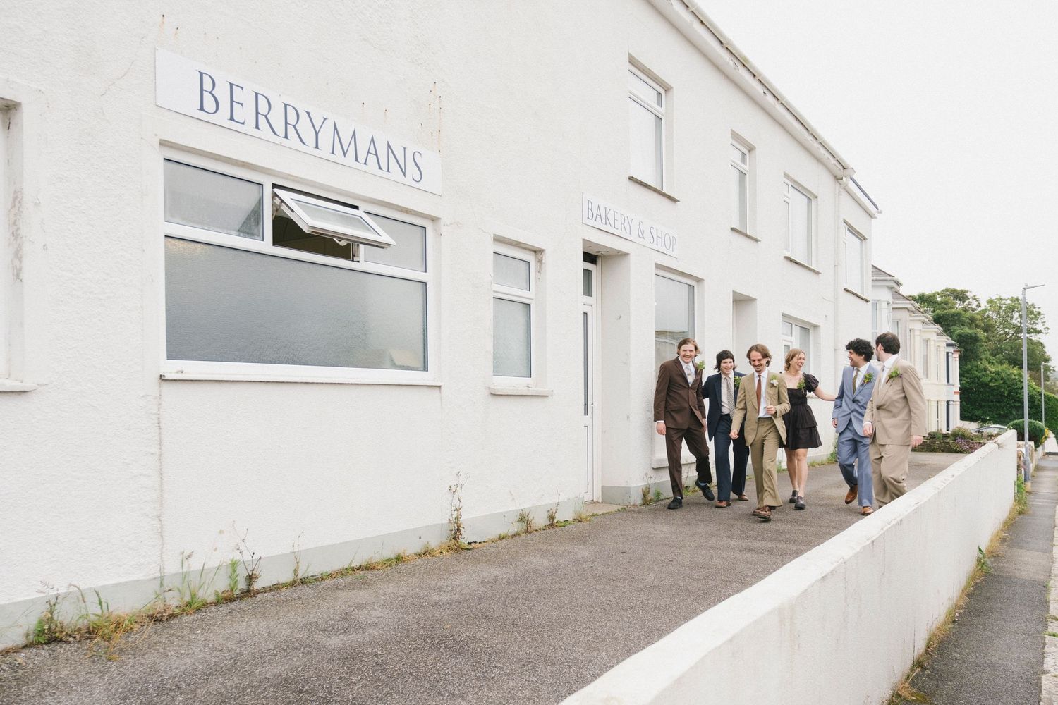 Wedding guests walking along a white building exterior with a Berrymans sign on an overcast day.