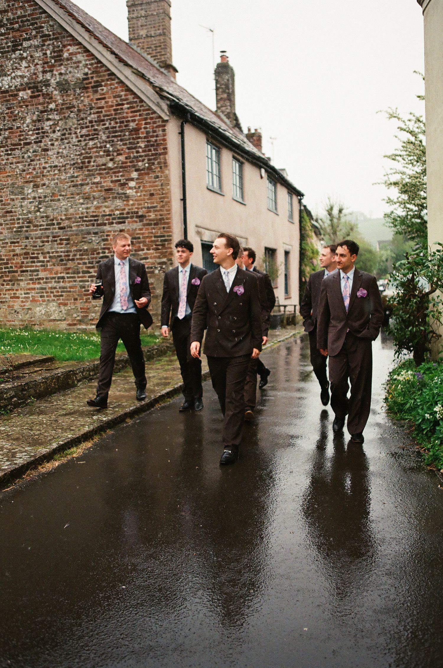 Groomsmen in dark suits with purple boutonnieres walking down a wet village street on a rainy wedding day.