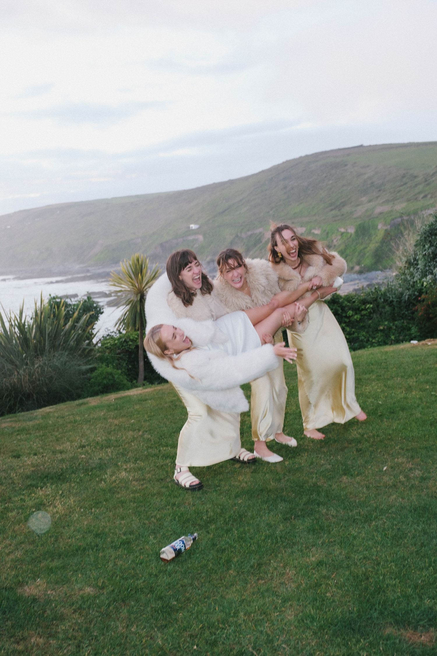Bridesmaids in yellow dresses playfully lifting bride on coastal lawn with ocean views behind.