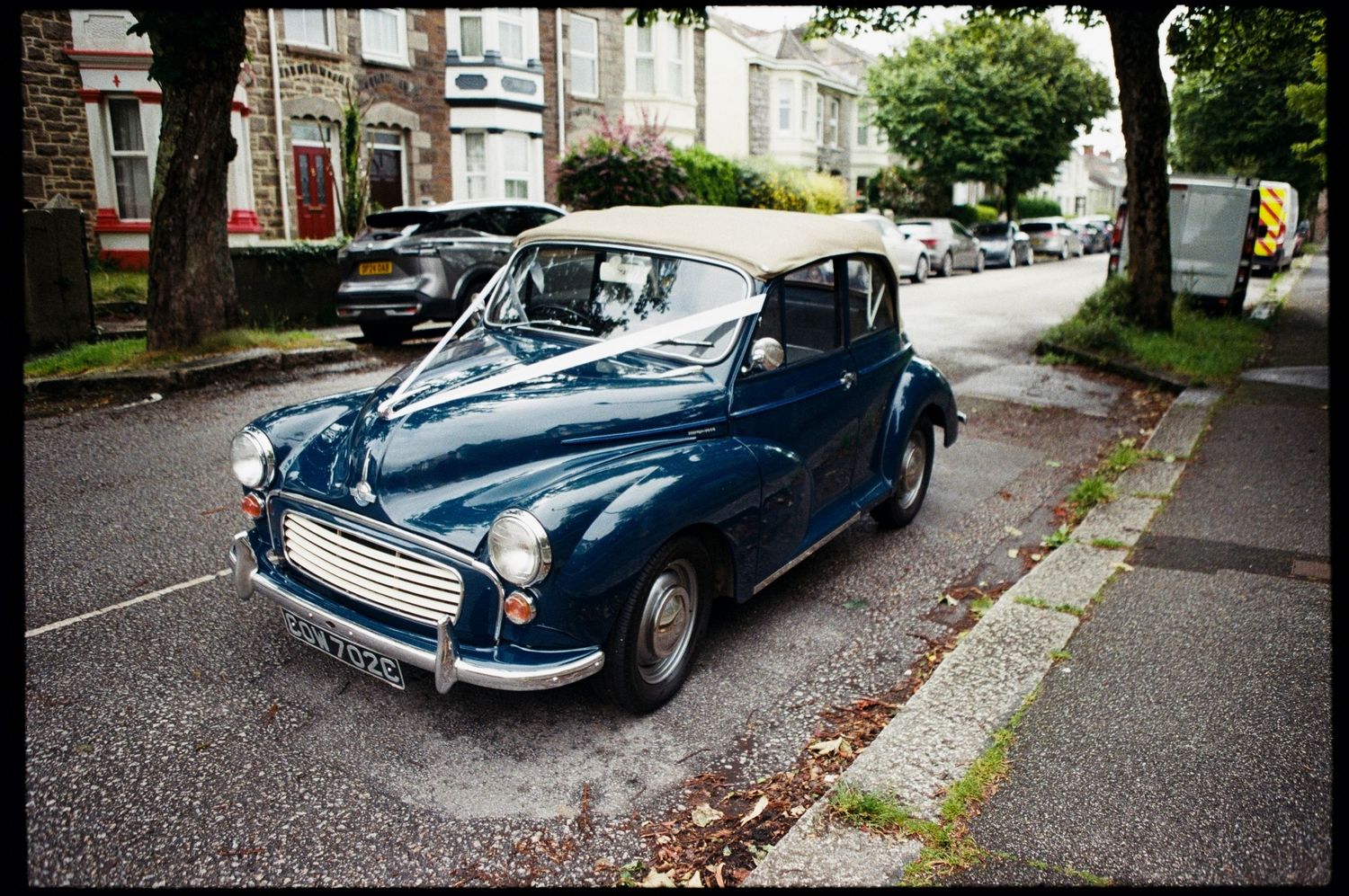 Vintage blue Morris Minor convertible parked on a quiet residential street, decorated with white ribbons.