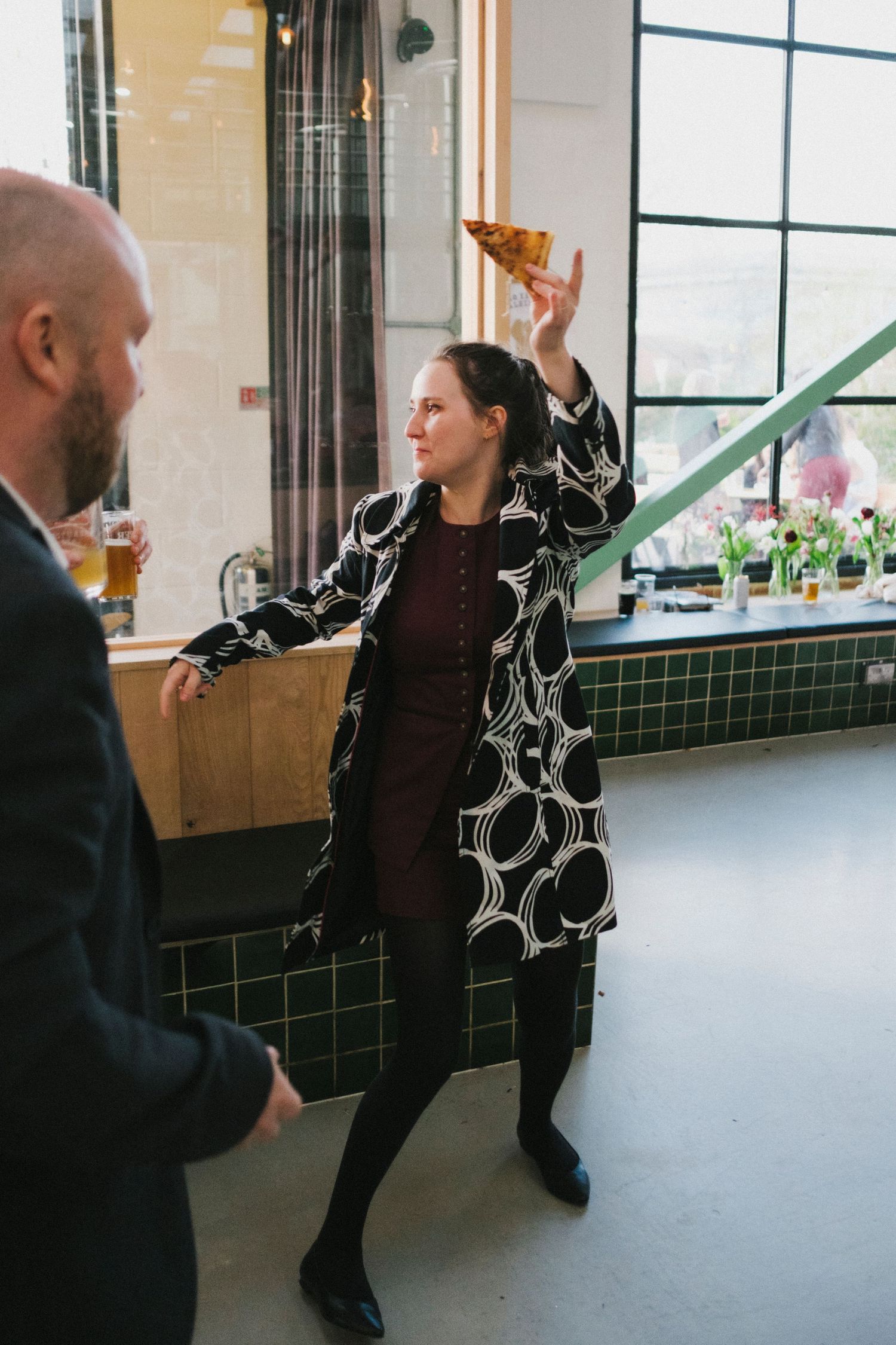 Wedding guest in bold patterned coat tossing pizza slice playfully at modern reception venue.