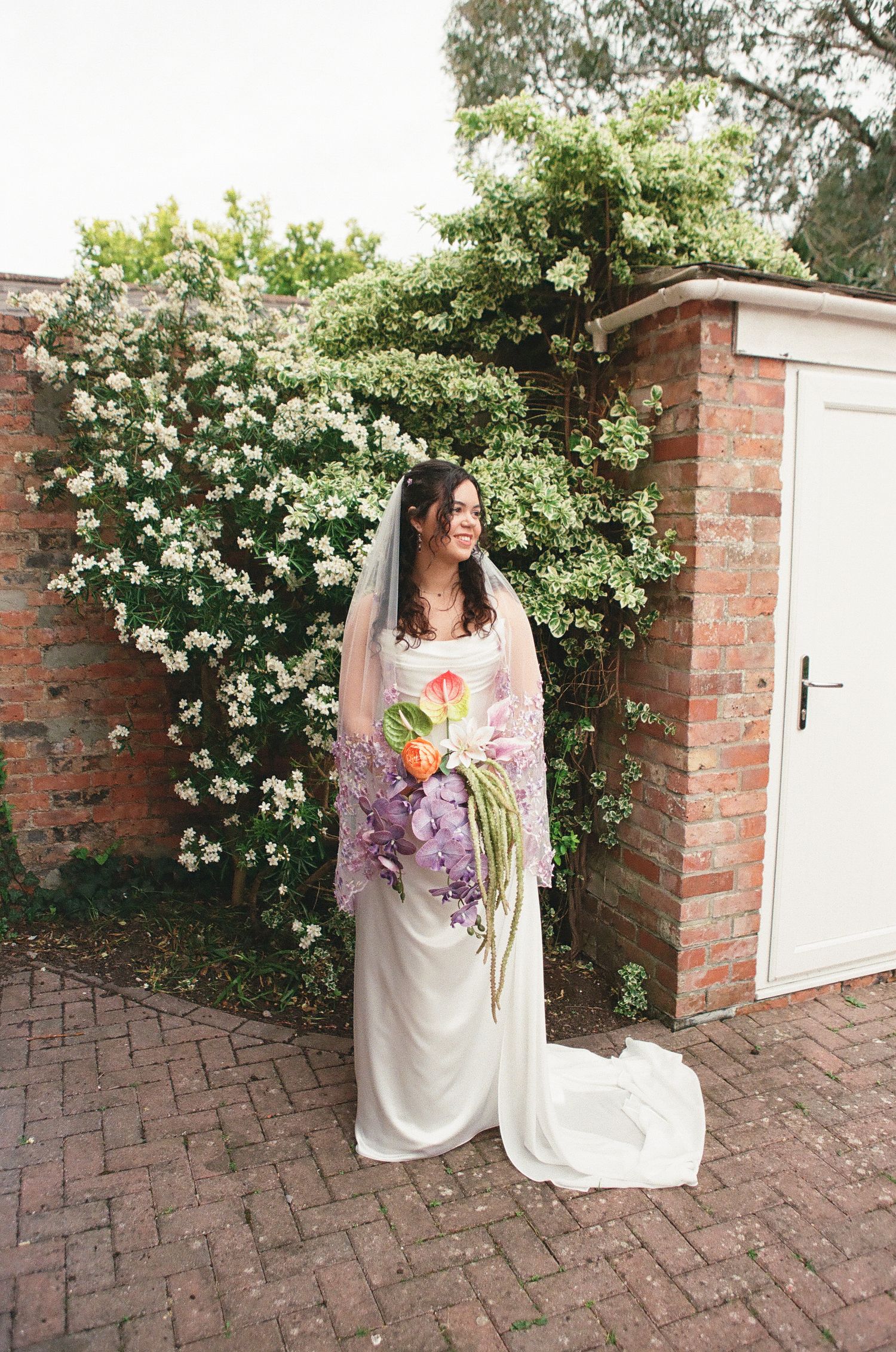 Bride in white gown holds a colorful bouquet standing before a flowering wall and brick garden.