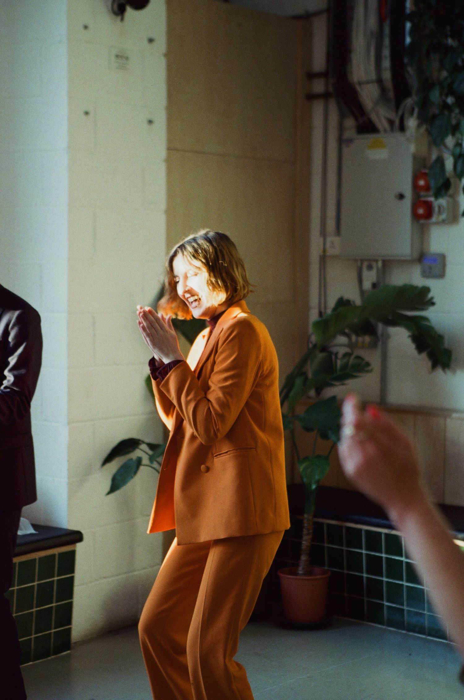 Woman in orange suit clapping hands and laughing in a tiled room with plants.