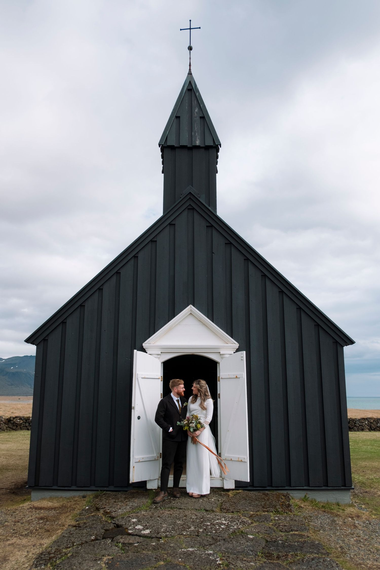 Bride and groom embracing at the entrance of a iconic black wooden church under a cloudy sky.