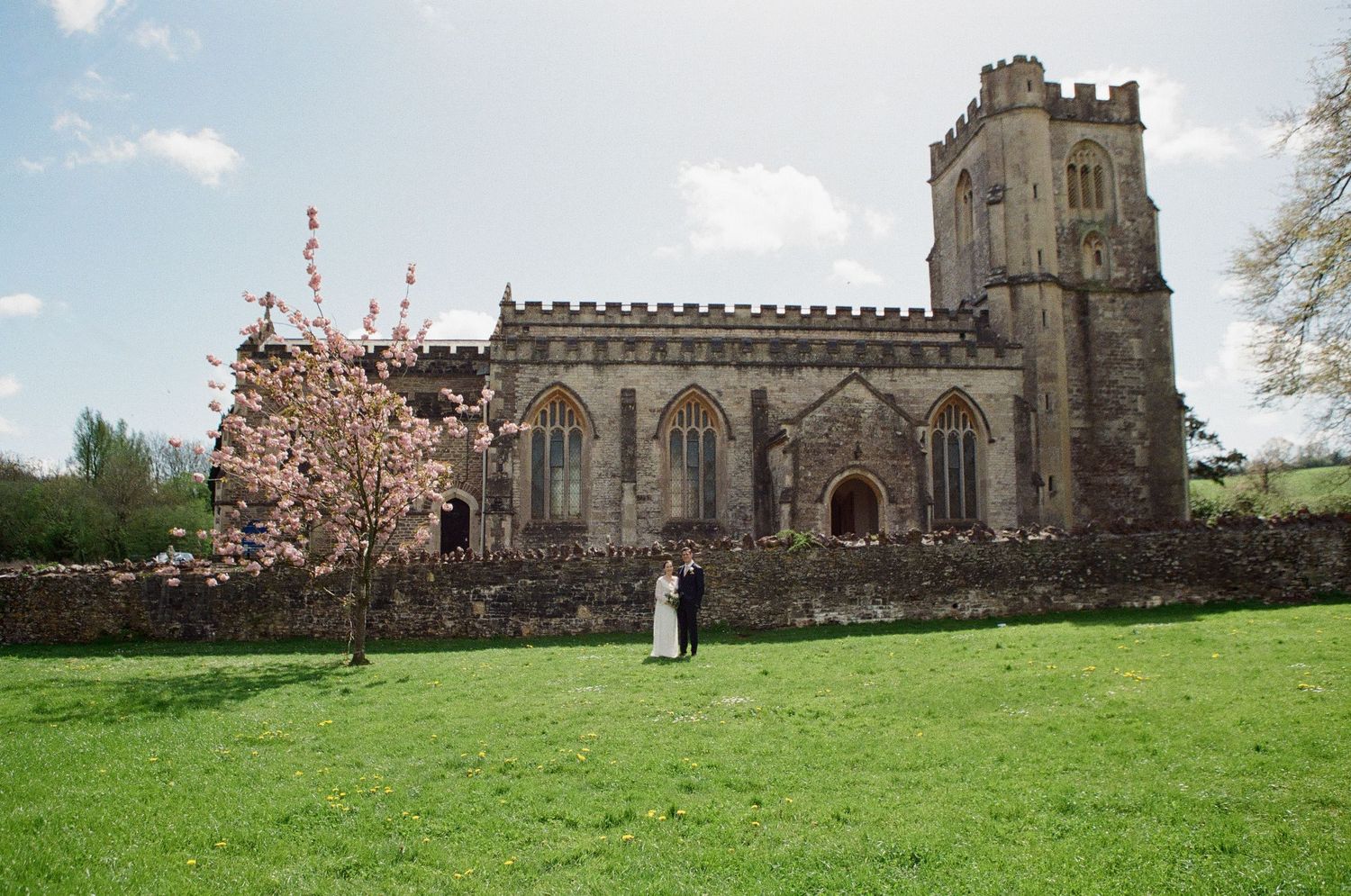 Bride and groom standing before grand historic English church with pink cherry blossom tree.