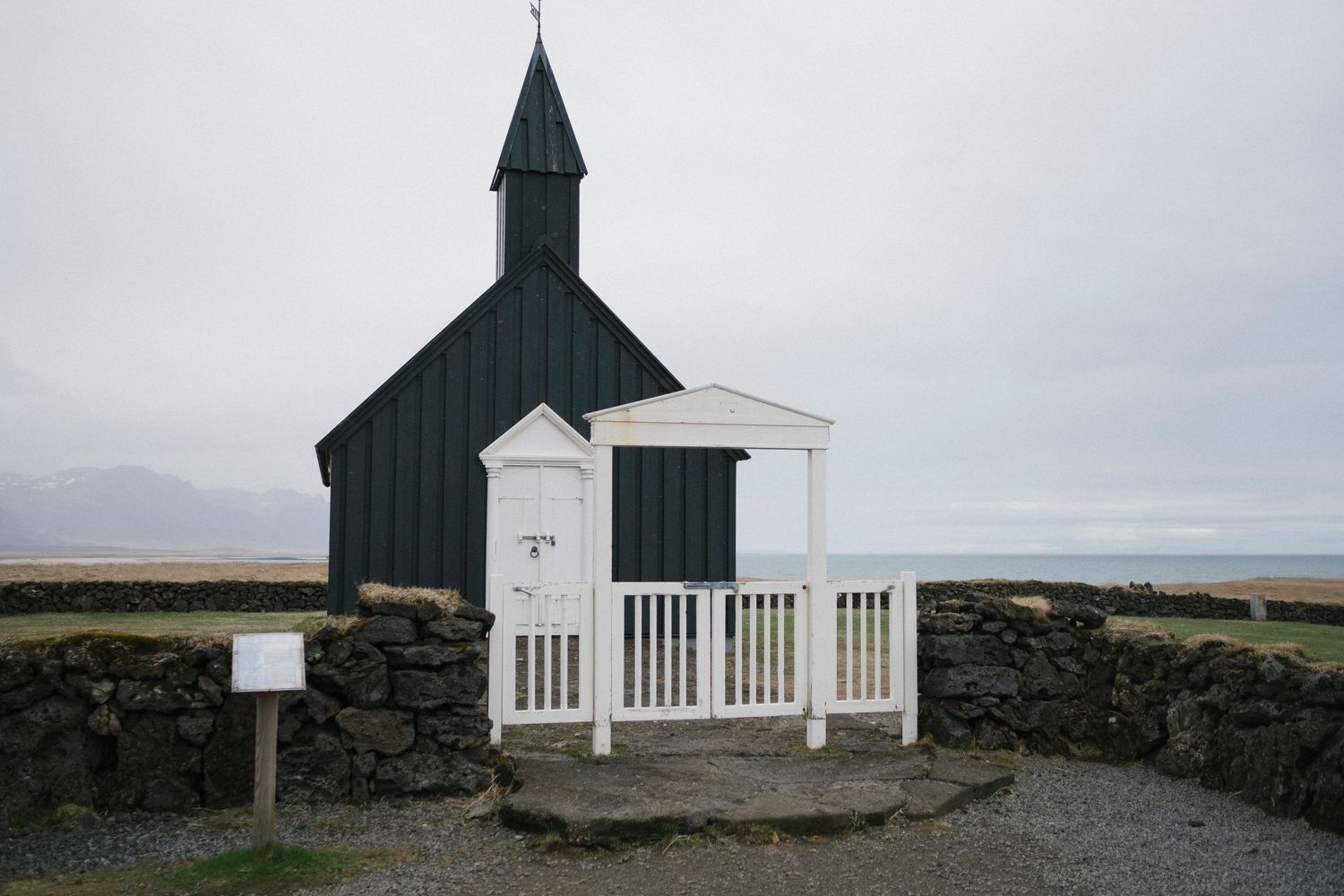 A black wooden church with a white gate and stone wall under an overcast sky in Iceland.