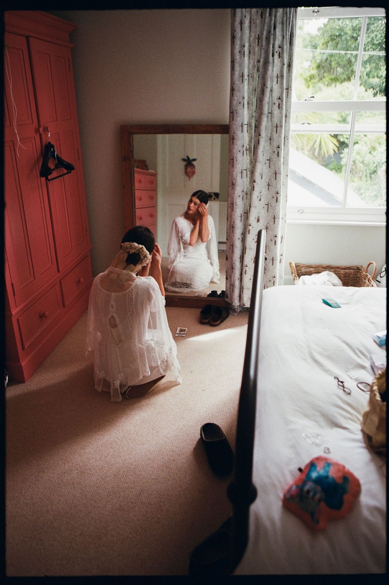 Bride in white wedding dress kneeling before mirror while getting ready in a pink bedroom.