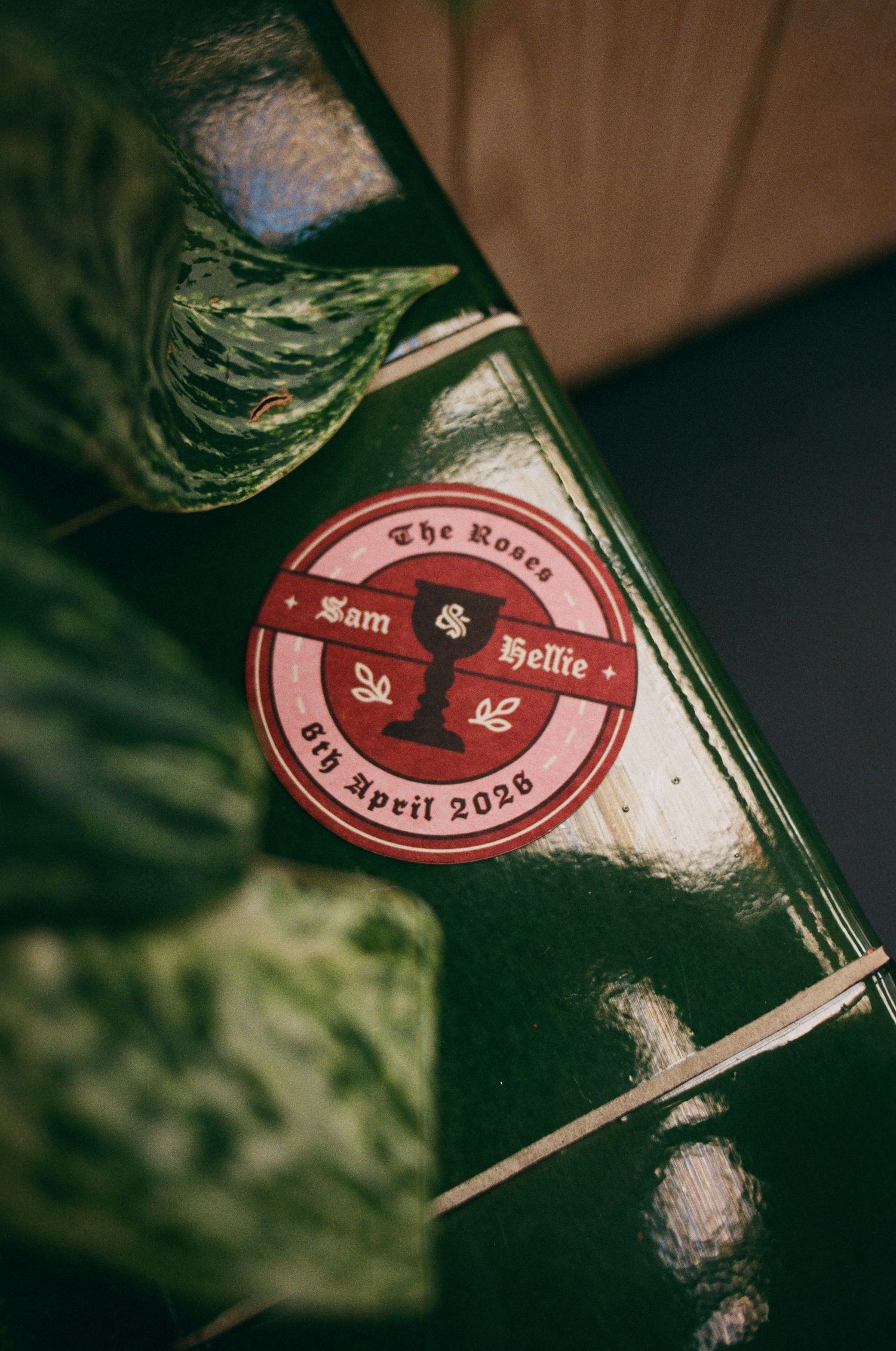 A circular red and dark wedding sticker reading The Roses, Sam and Hellie, 6th April 2026, placed on a green surface.