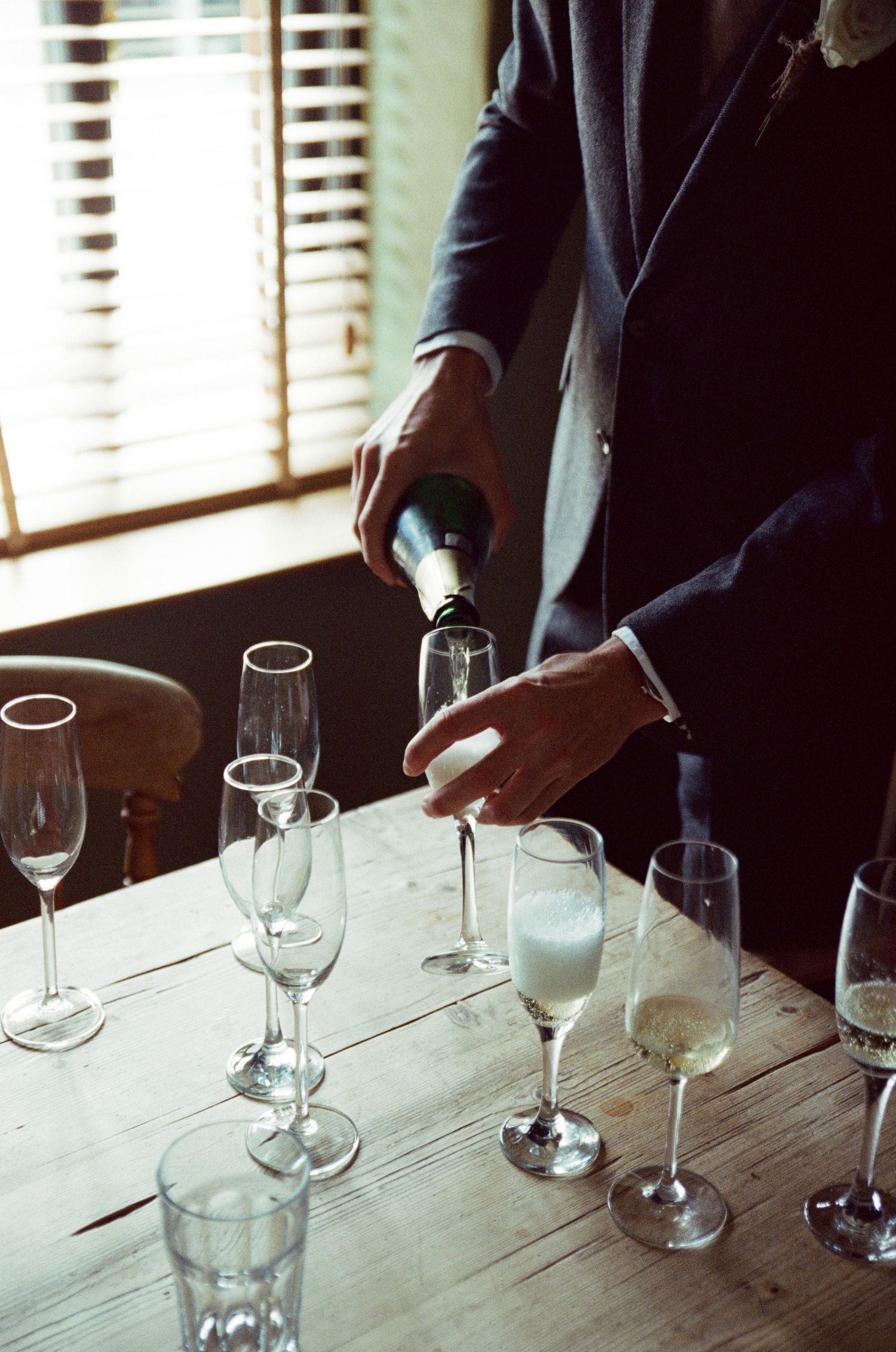 Man in suit pouring champagne into flute glasses on rustic wooden table near window with blinds.