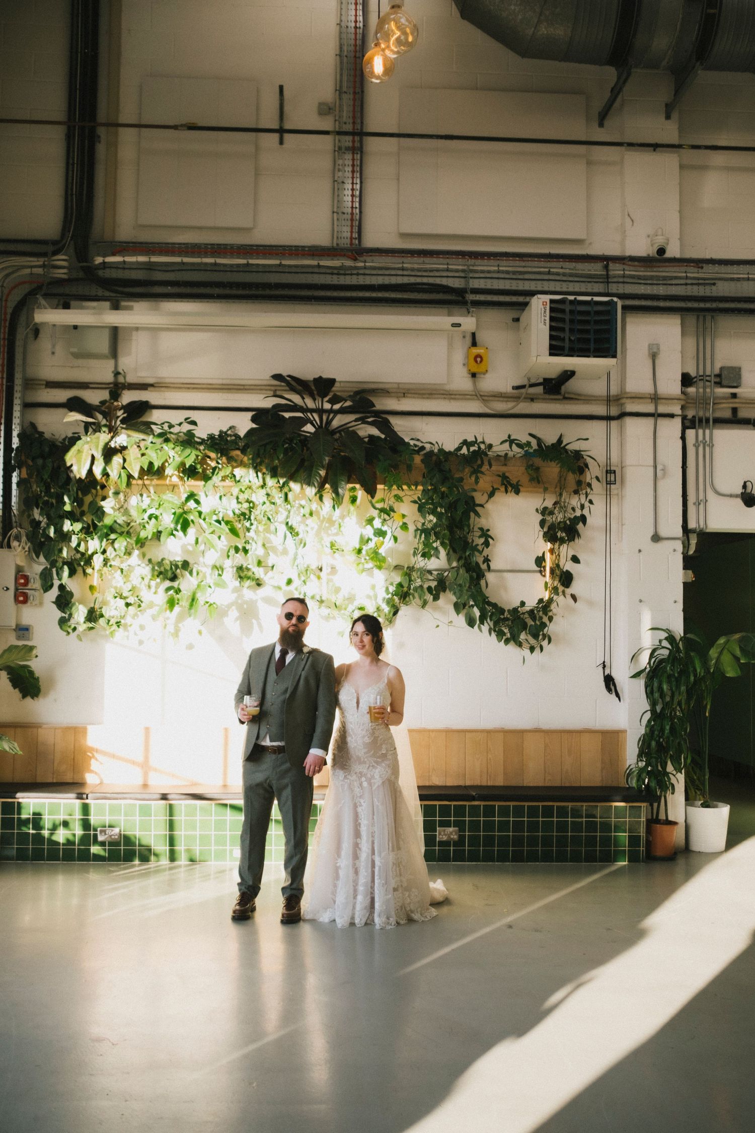 Bride and groom stand together holding drinks beneath a lush hanging plant installation in an industrial wedding venue.