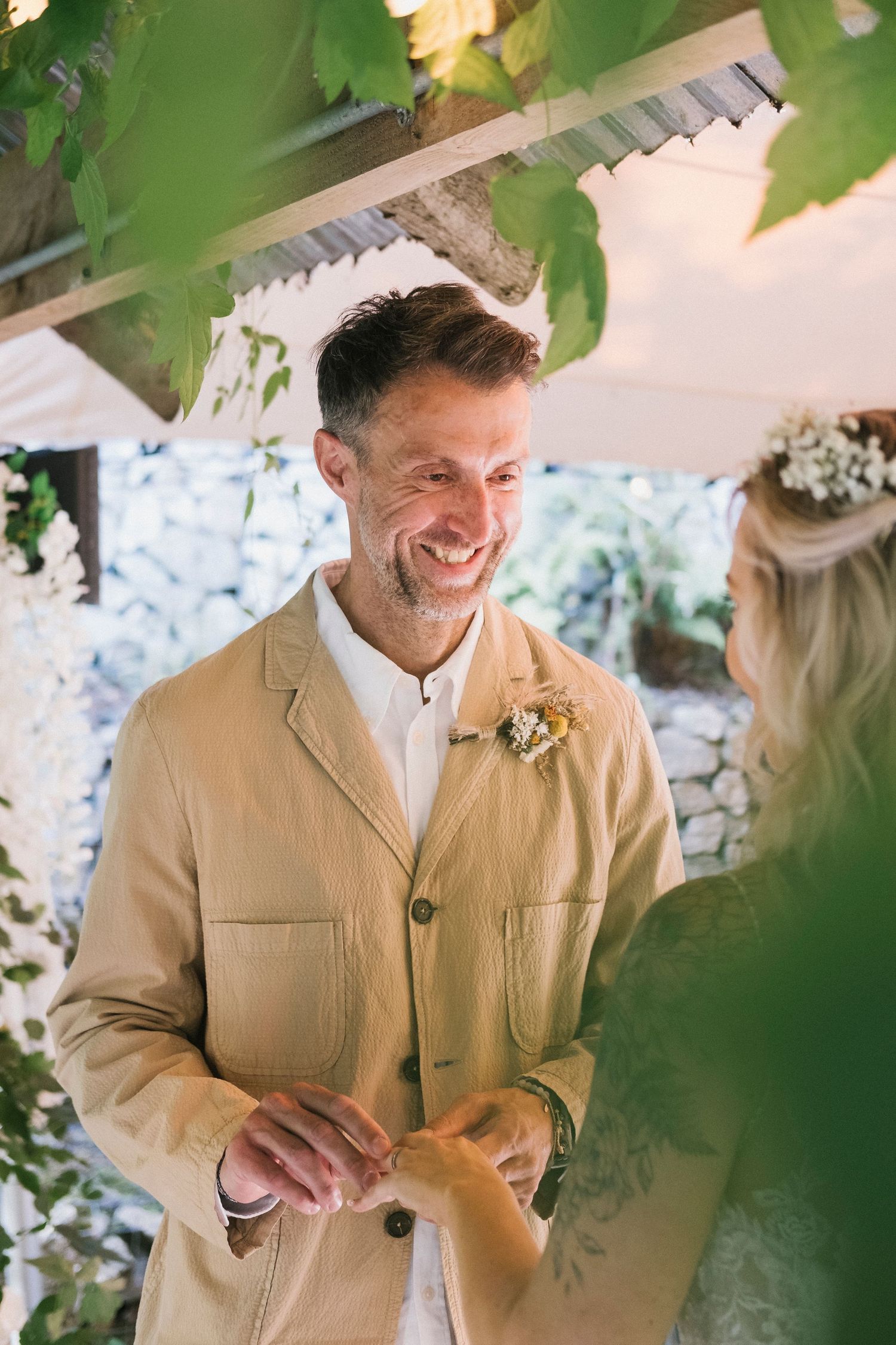 Smiling groom in a beige jacket exchanges rings with his bride during an outdoor ceremony.