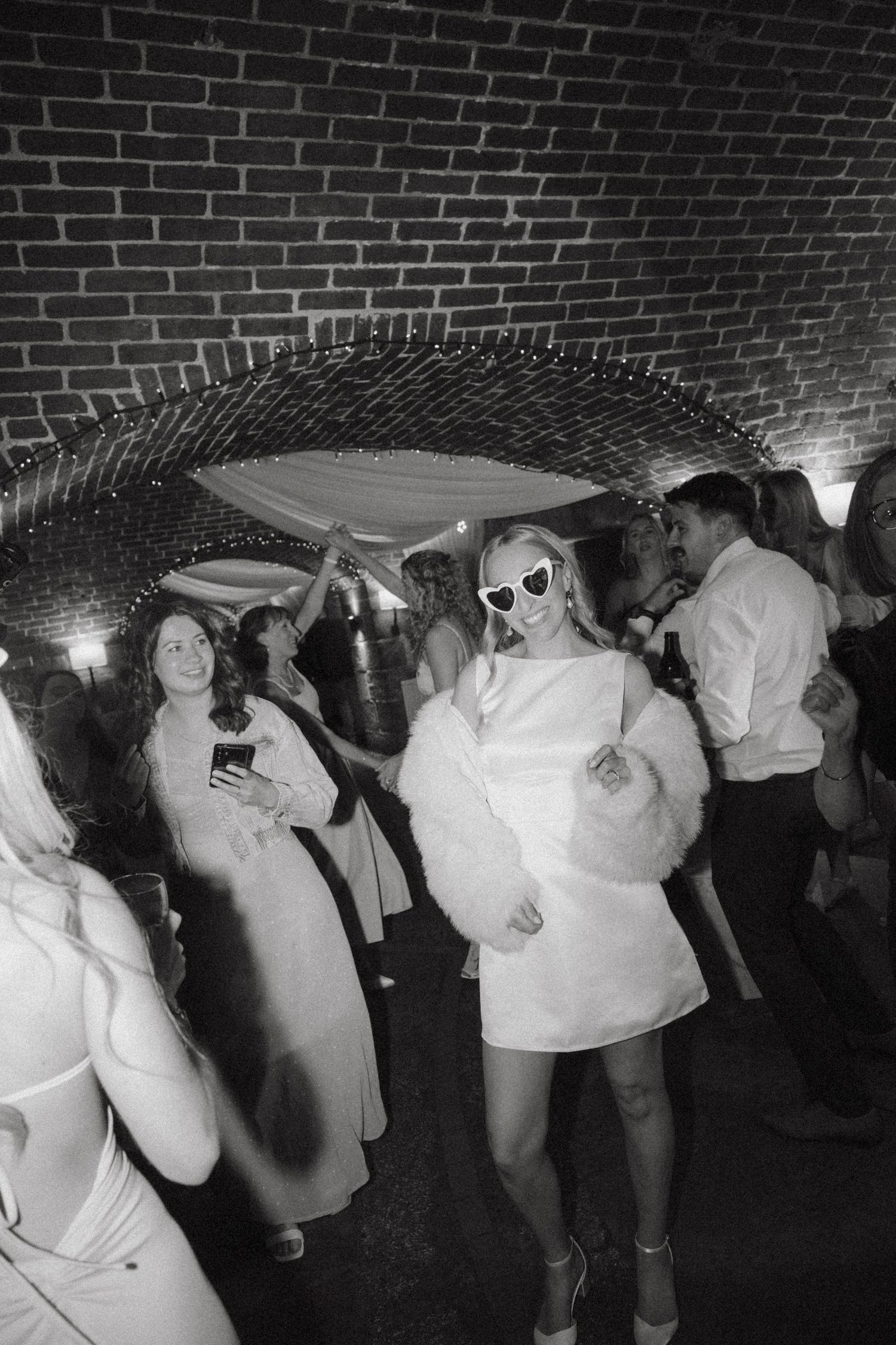 Bride in white mini dress and fur coat dancing under disco ball at wedding reception party.