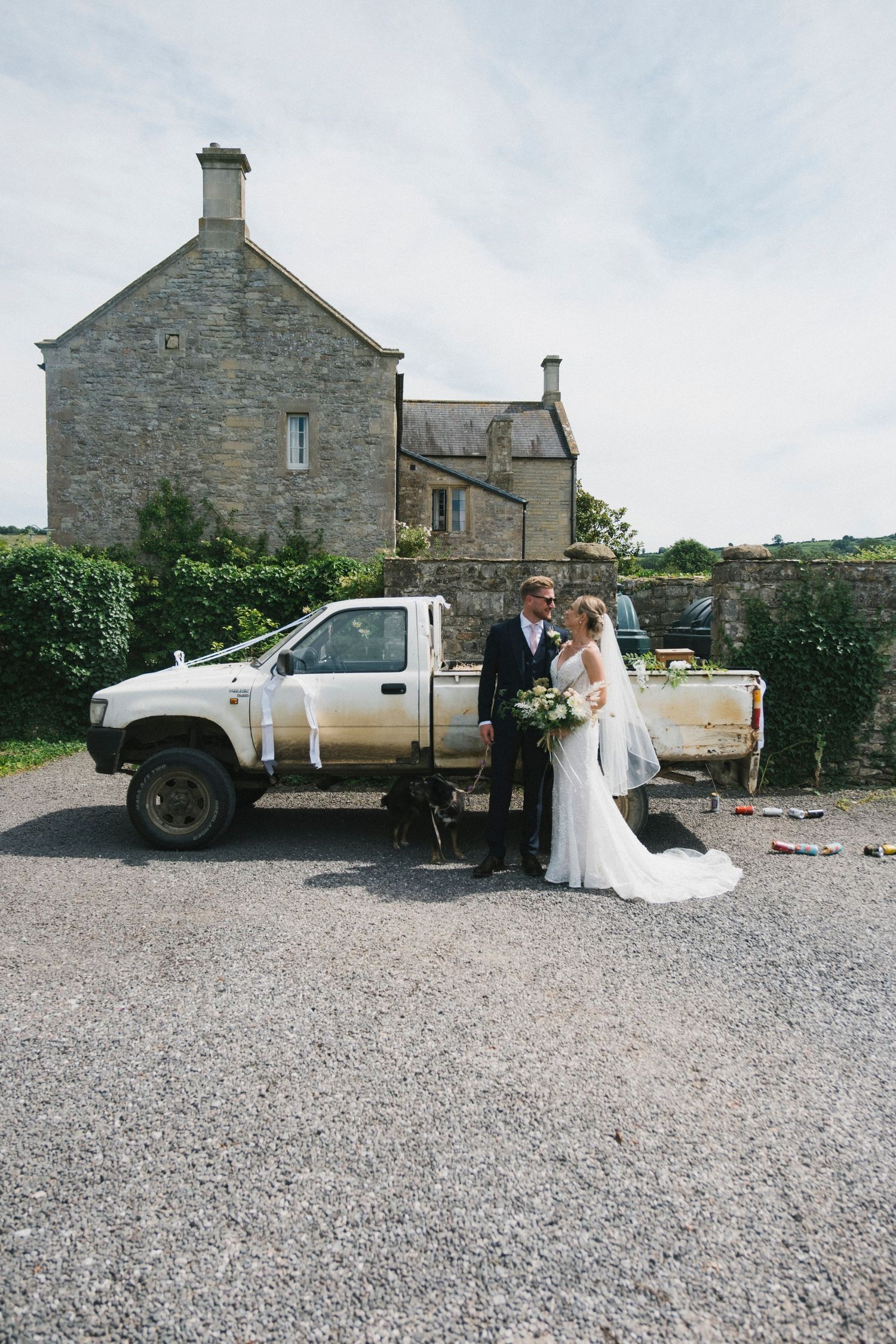 Bride and groom kissing beside a decorated pickup truck in front of a rustic stone farmhouse on their wedding day.