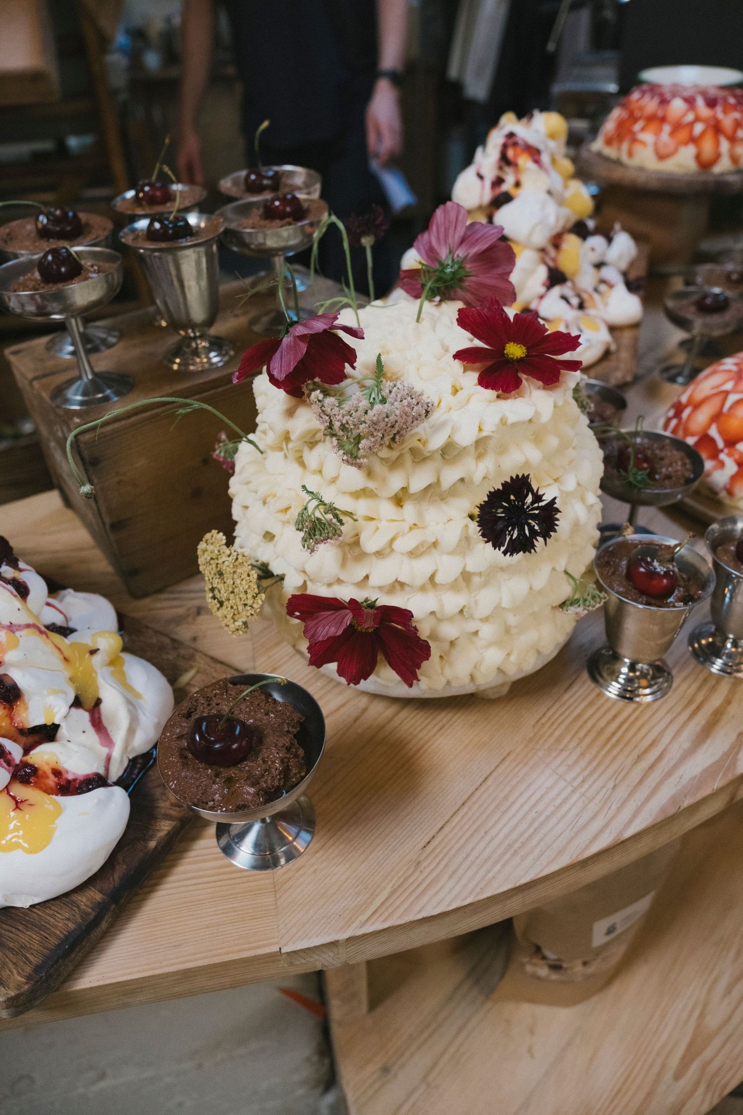 Wedding cake decorated with fresh flowers surrounded by assorted desserts on a wooden table.