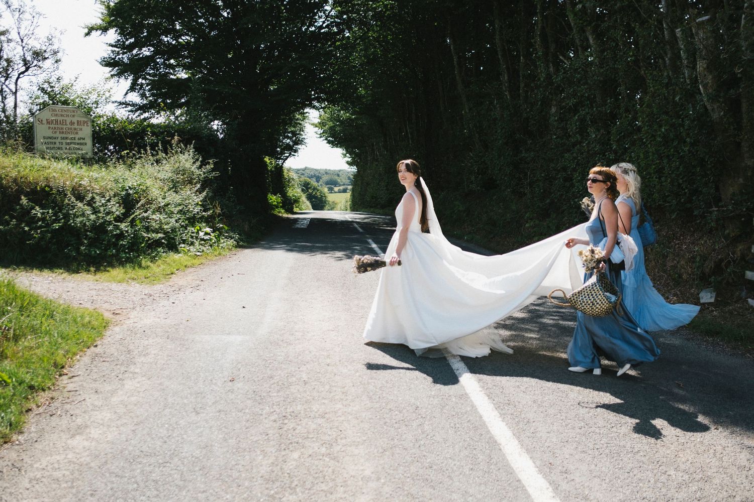 Bride in white gown crosses a country road with bridesmaids in blue dresses on a sunny day.