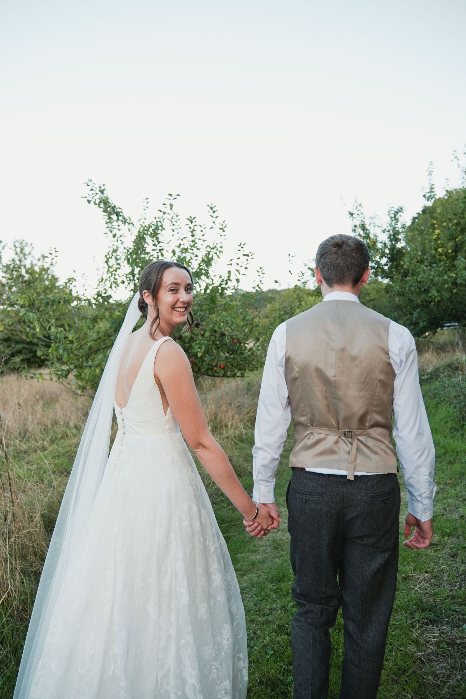 Bride in white gown and veil holds hands with groom in vest, walking through green meadow, smiling back at camera.