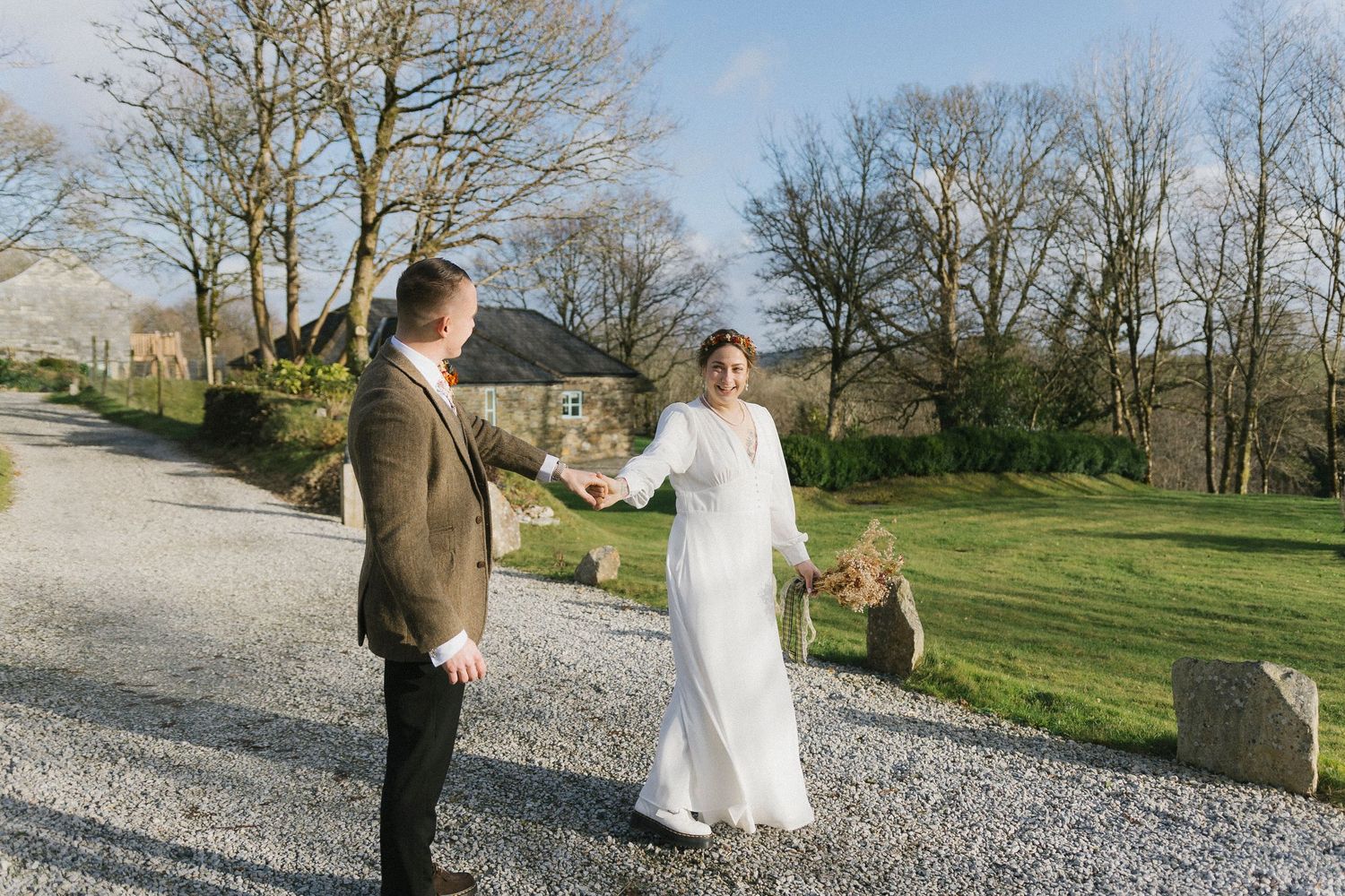 Bride in a white gown holds hands with her groom on a sunlit gravel driveway.