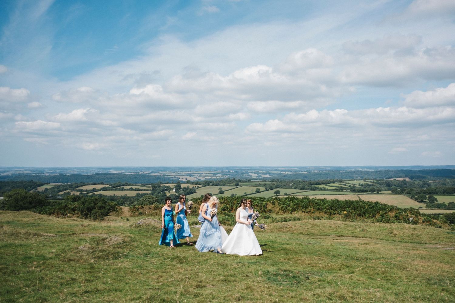 Bride and bridesmaids in blue dresses walk across a hilltop with a sweeping countryside view.