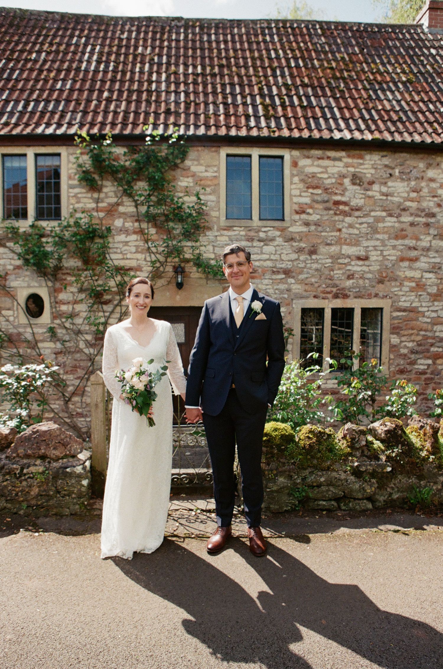 Bride in elegant white gown and groom in navy suit pose together outside a rustic stone farmhouse.