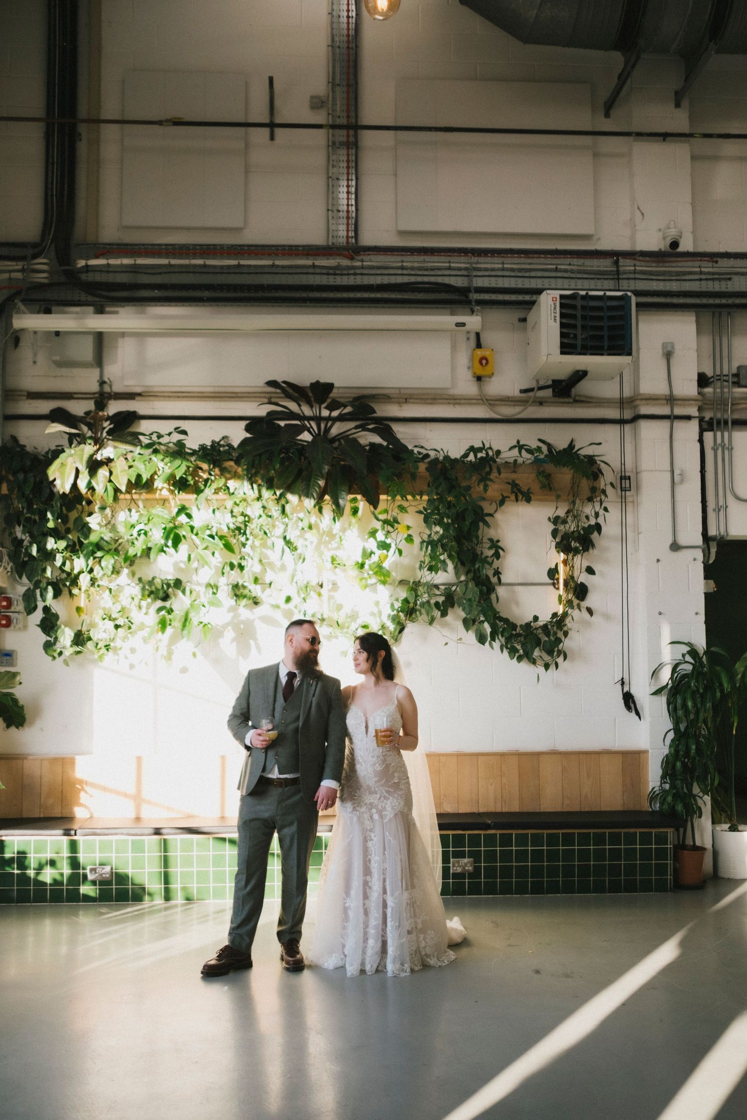 Bride and groom sharing intimate moment under lush green floral installation in industrial wedding venue.