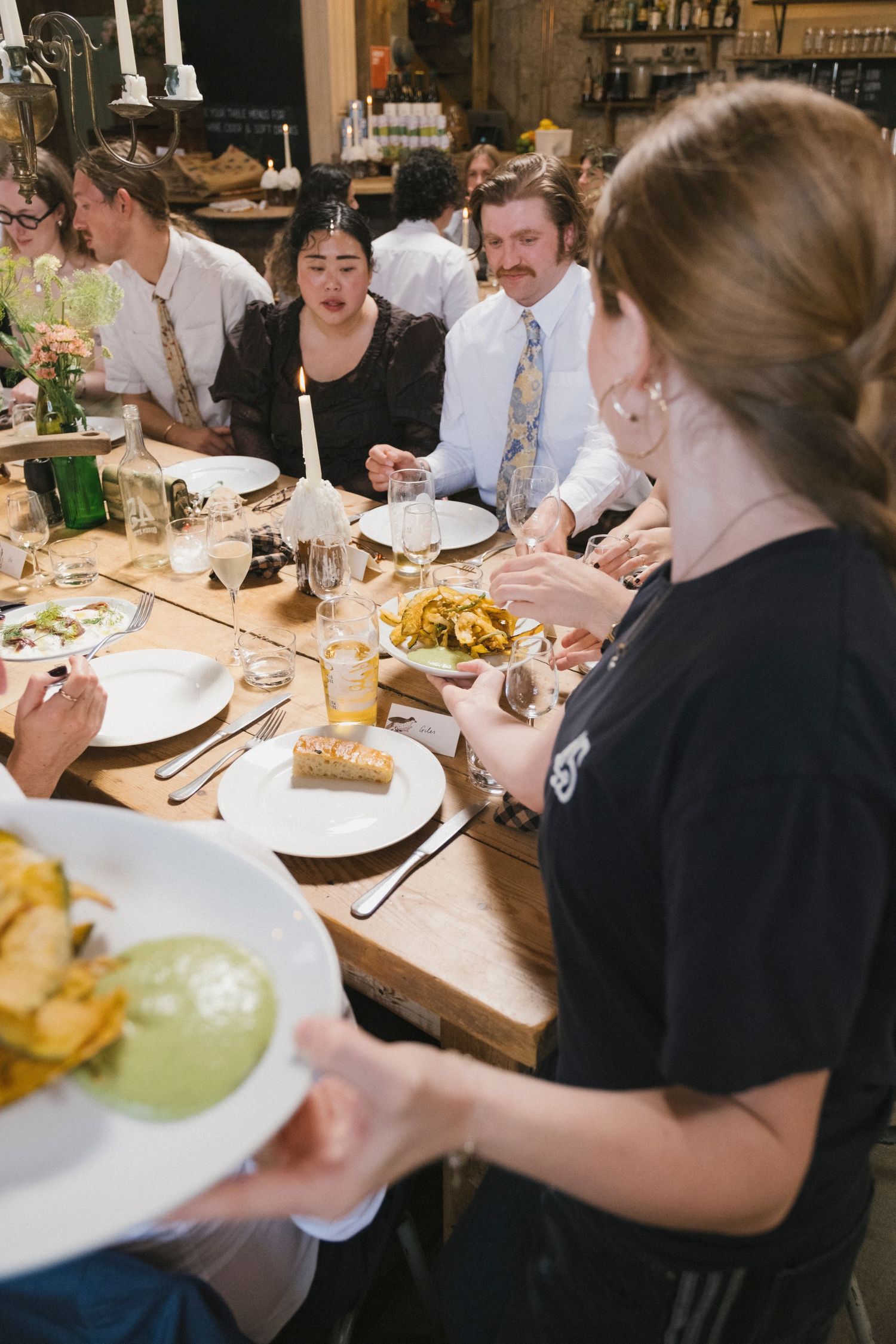 A server delivers plates of food to elegantly dressed guests dining at a candlelit wedding reception table.