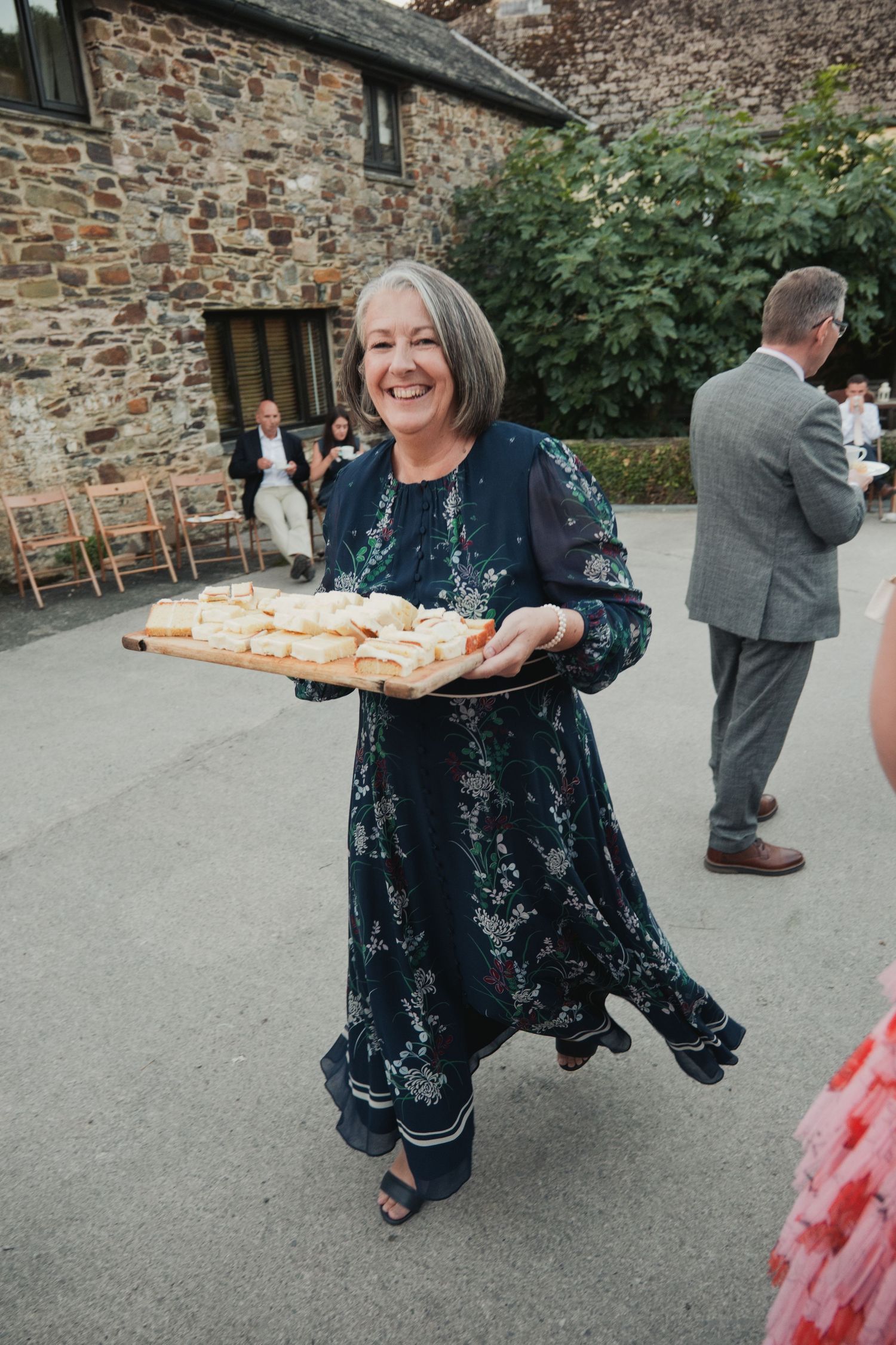 Smiling woman in floral dress carrying tray of sandwiches at outdoor wedding reception near stone barn.