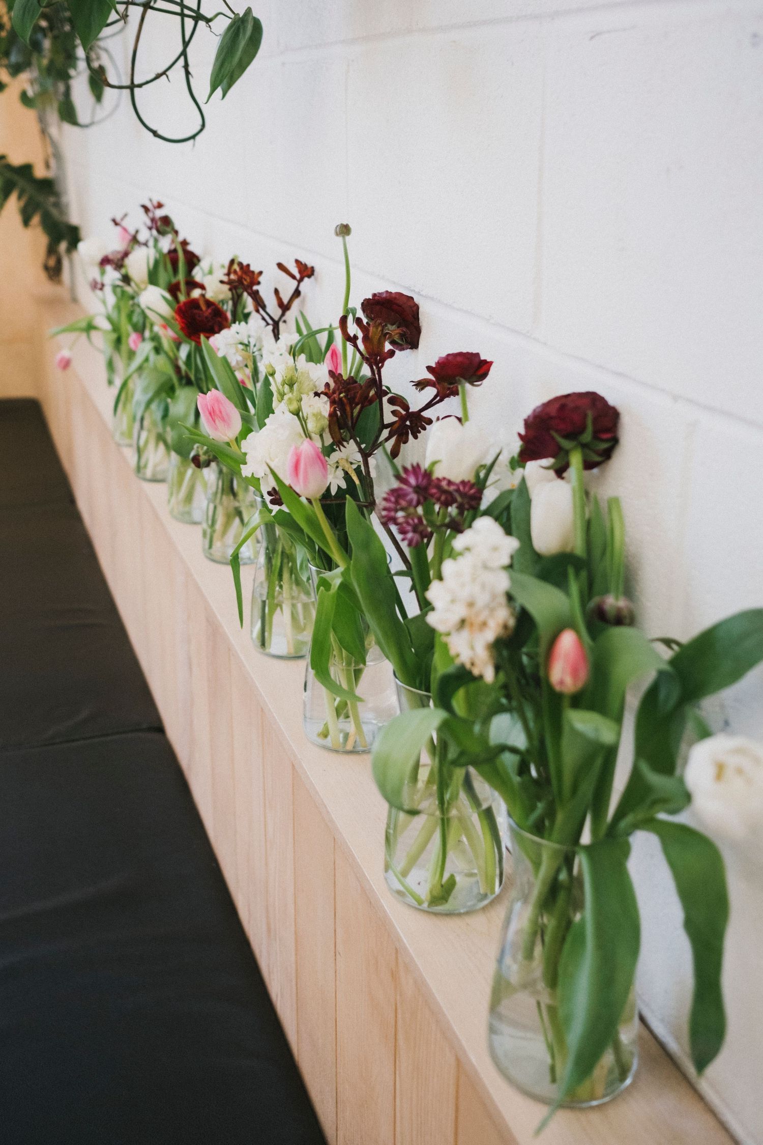 Row of glass vases with pink, white, and burgundy flowers on wooden shelf against white brick wall.